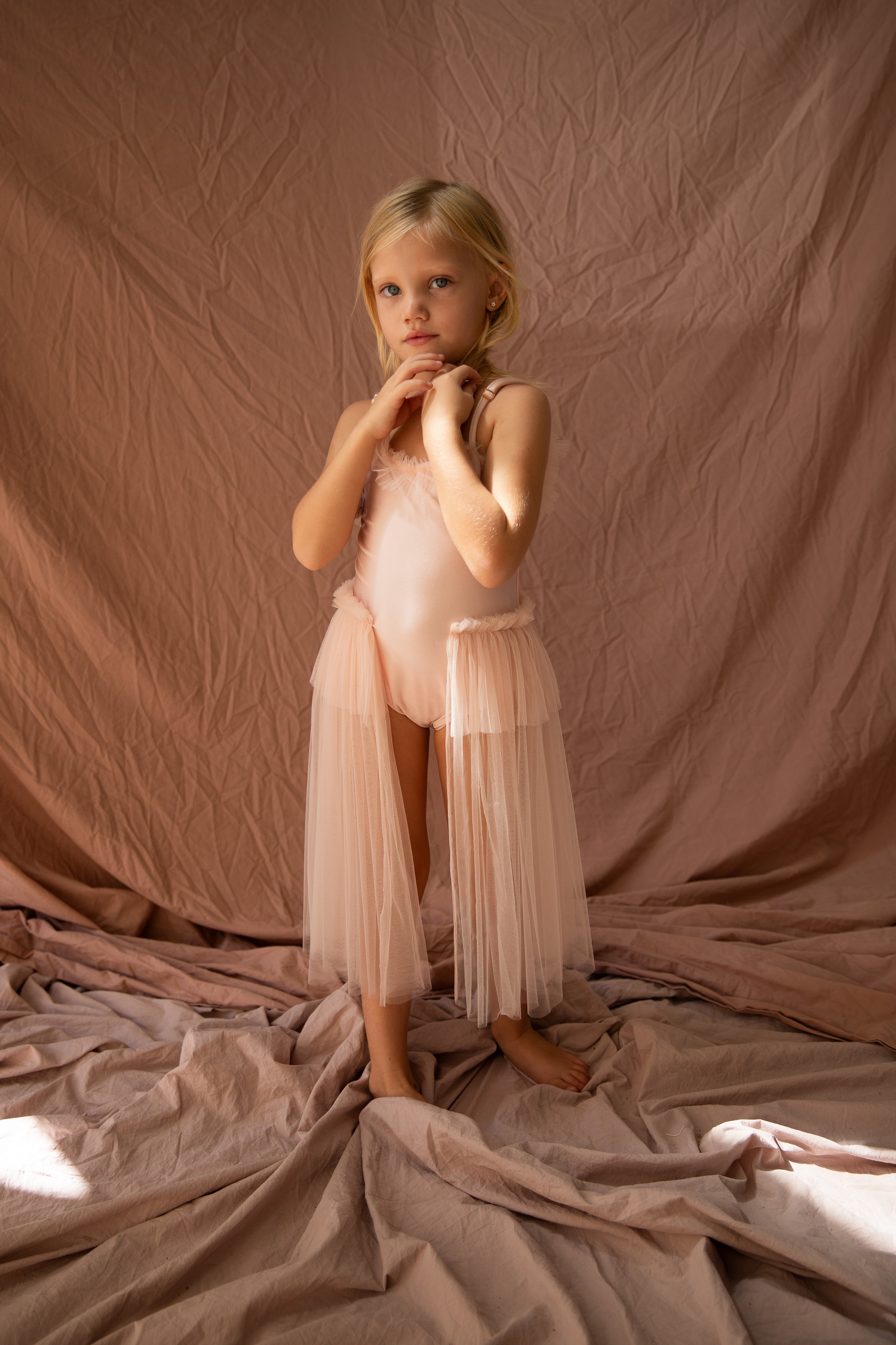 Elanore, a young girl with blonde hair, stands barefoot on a beige fabric backdrop. She is dressed in the BELLA + LACE Elanore Dress Twinkle Toes - a light pink, flowing dress featuring tulle details reminiscent of a ballet class outfit. With her hands clasped near her chin, Elanore adopts a thoughtful expression.