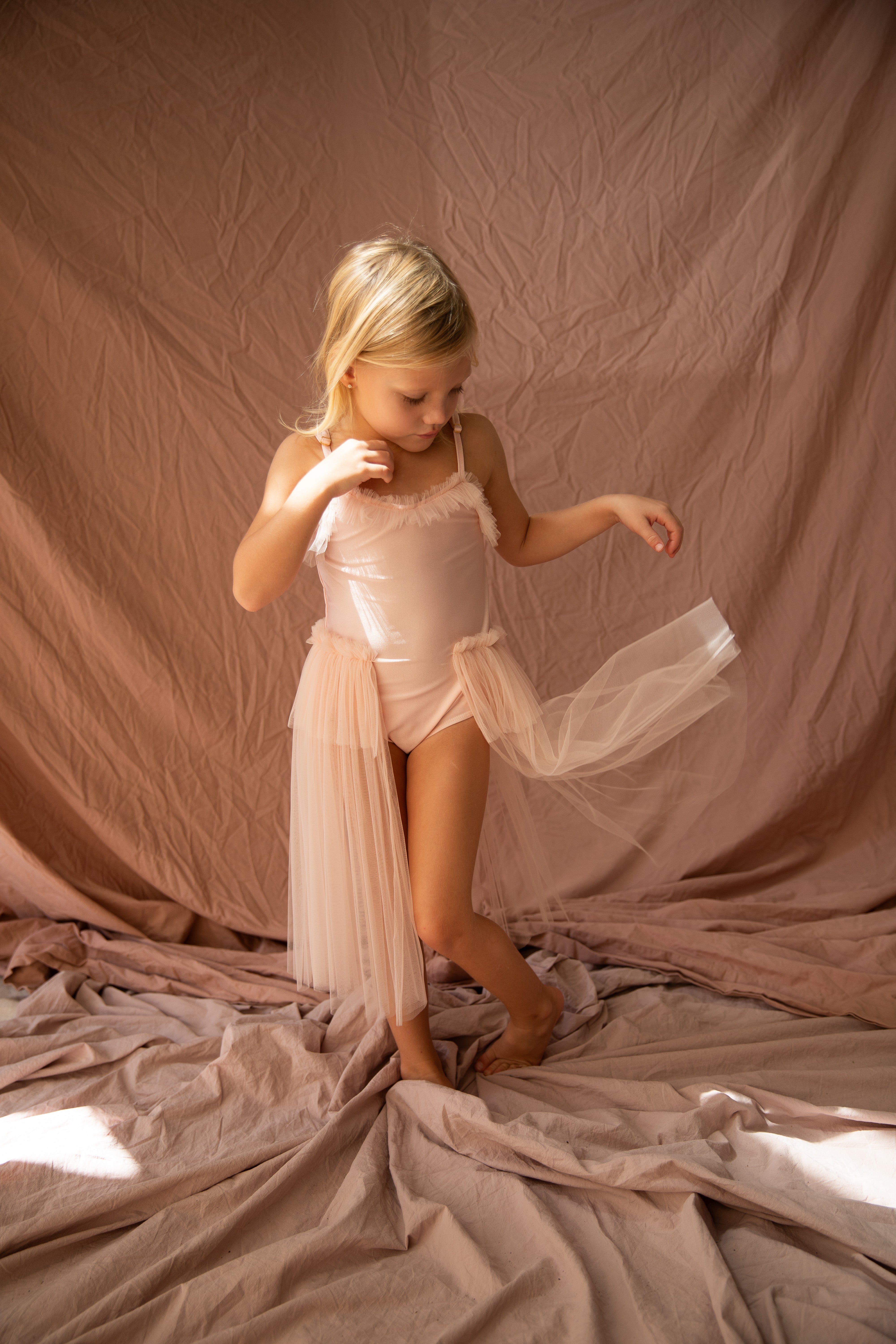 A young child with blonde hair, reminiscent of Elanore from ballet class, is wearing the BELLA + LACE Elanore Dress Twinkle Toes in light pink tulle. She seems to be dancing or twirling on a softly lit, fabric-covered floor with a matching fabric backdrop.