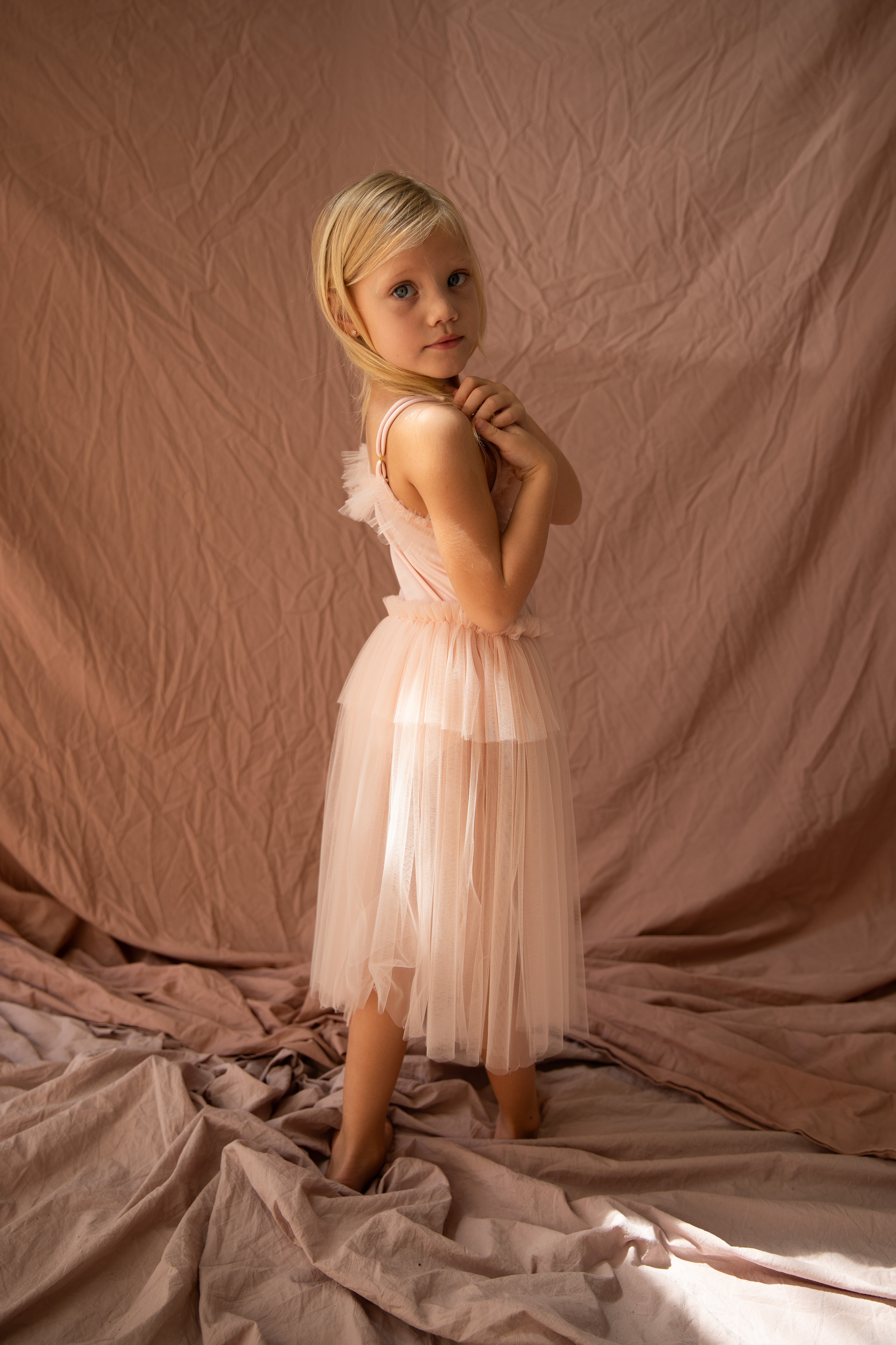 A young girl with blonde hair, Elanore, stands gracefully in the Elanore Dress Twinkle Toes by BELLA + LACE, a pale pink tutu dress crafted from delicate tulle. She poses with hands folded near her neck against a soft pink fabric backdrop, evoking the serene elegance of a ballet class.