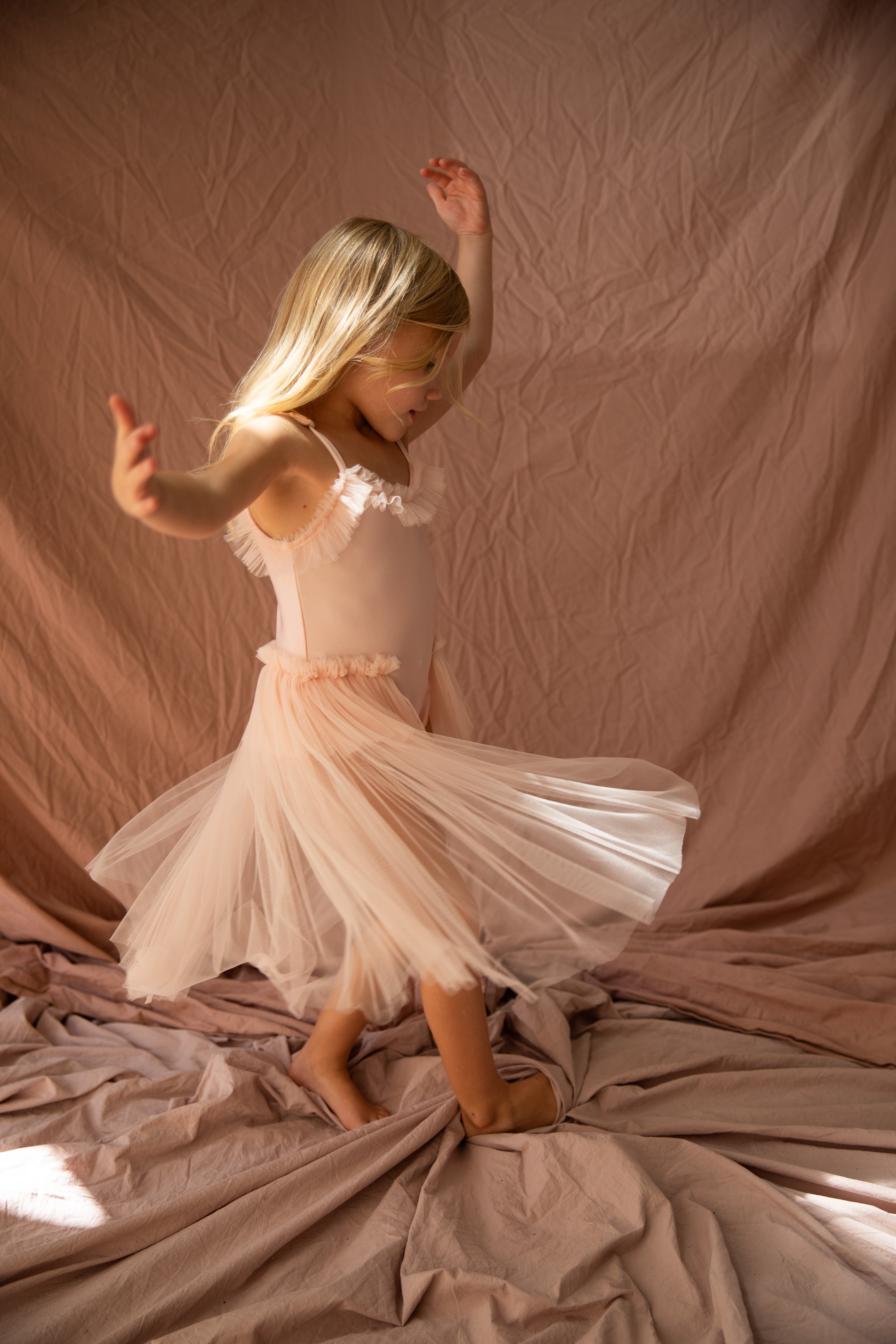 A young girl named Elanore, with long blonde hair, is twirling in the Elanore Dress Twinkle Toes by BELLA + LACE. She is barefoot, standing on soft, crumpled fabric in a warm, softly lit setting reminiscent of a ballet class.