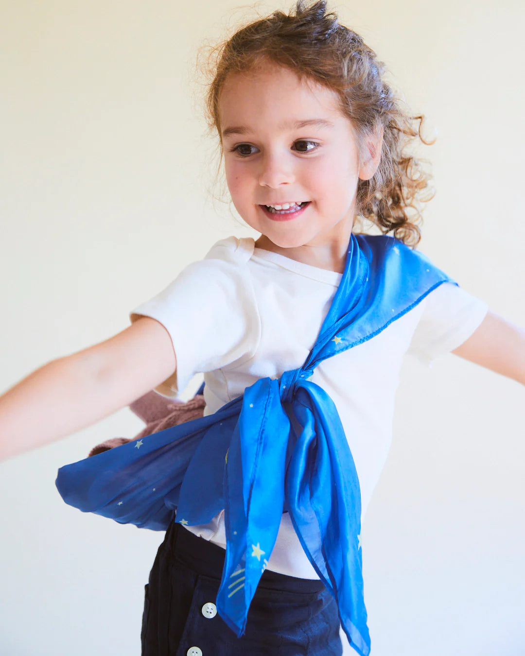 A young child with curly hair smiles while wearing a white T-shirt and dark pants with buttons. Around their neck is an Enchanted Playsilk Starry Night by SARAH'S SILKS, tied like a cape and offering open-ended play against the light background.