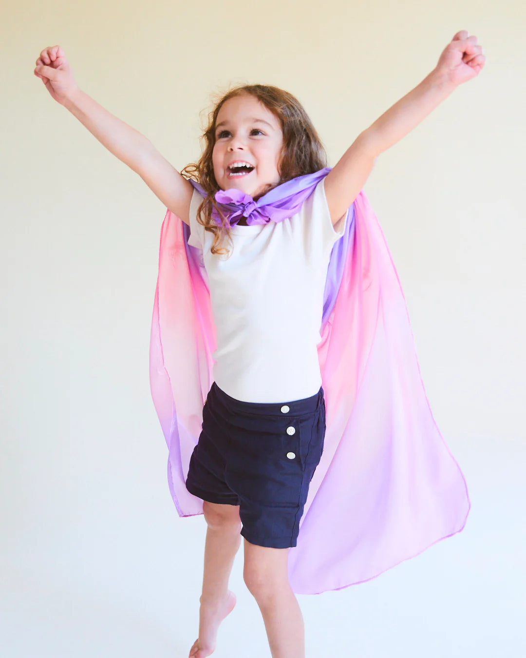 A joyful child with curly hair, wearing an Enchanted Playsilk Blossom cape by SARAH'S SILKS in pink and purple, pairs it with a white shirt and dark shorts. They leap with arms raised, smiling against a light background—a perfect moment of open-ended play that unlocks their creative potential.