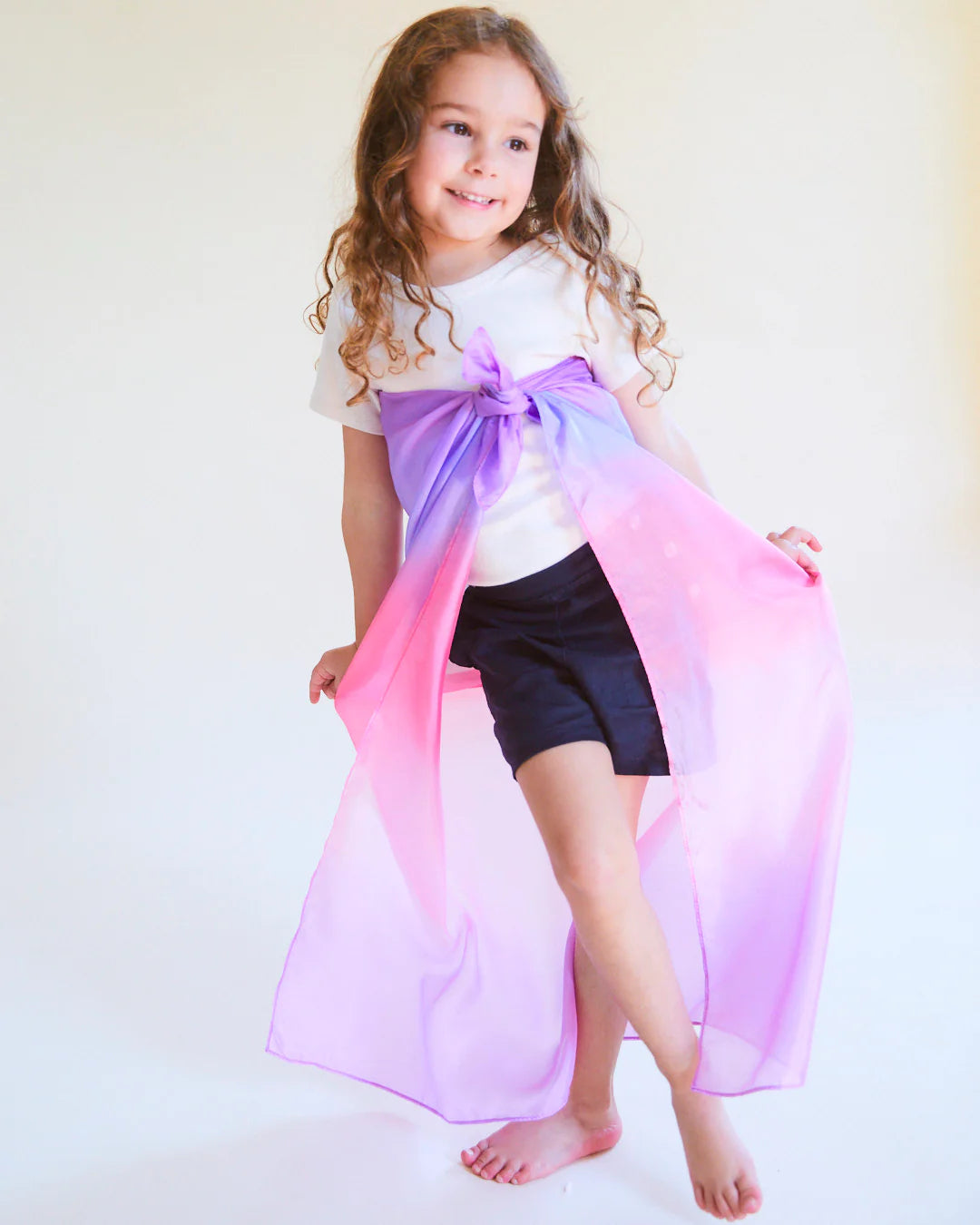 A young girl with curly hair, dressed in a white shirt and dark shorts, stands barefoot and smiling against a simple light background. She joyfully wraps an Enchanted Playsilk Blossom from SARAH'S SILKS—a sheer fabric with a purple and pink ombre design—around herself, inviting open-ended play and inspiring her creativity.