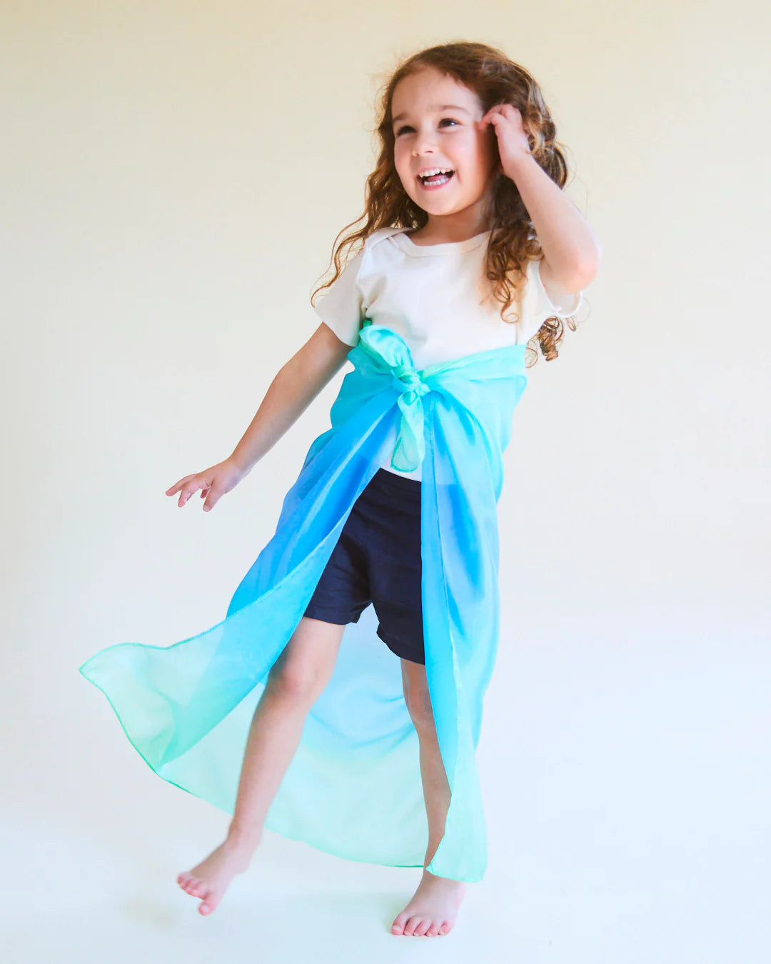 A young child with curly hair stands barefoot in a white shirt and dark shorts, joyfully wearing the Enchanted Playsilk Sea from SARAH'S SILKS tied around their waist like a skirt. Smiling and touching their hair against a plain light background, they embody boundless creative potential through open-ended play.