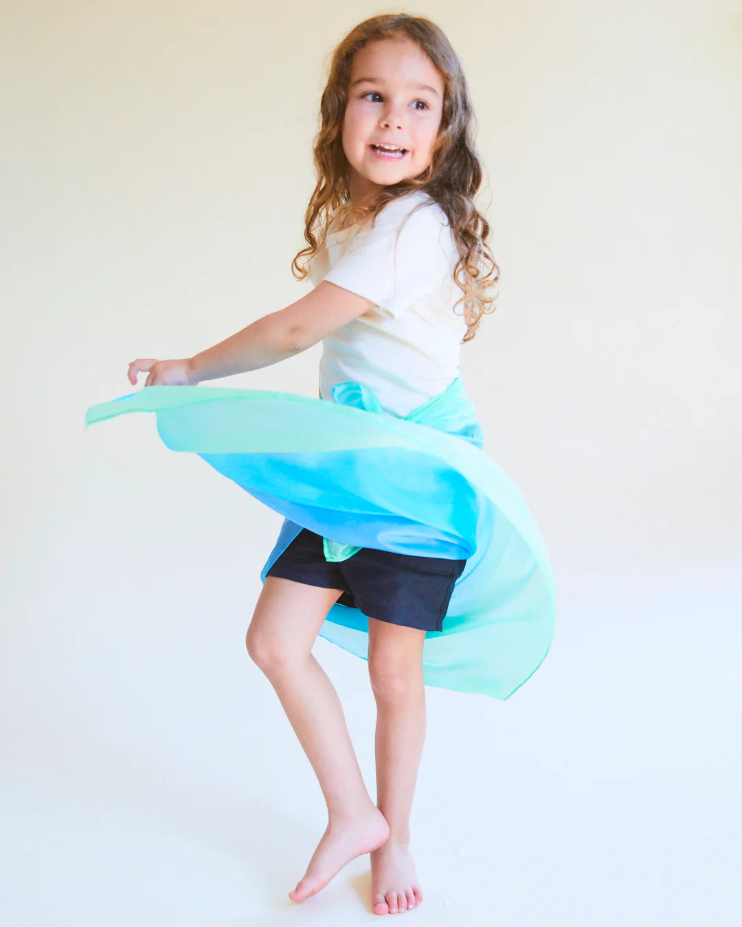 A young girl with long curly hair twirls joyfully, wearing a white top and a flowing Enchanted Playsilk Sea from SARAH'S SILKS as a skirt. She is barefoot, embracing the creative potential of open-ended play against a soft, neutral background.