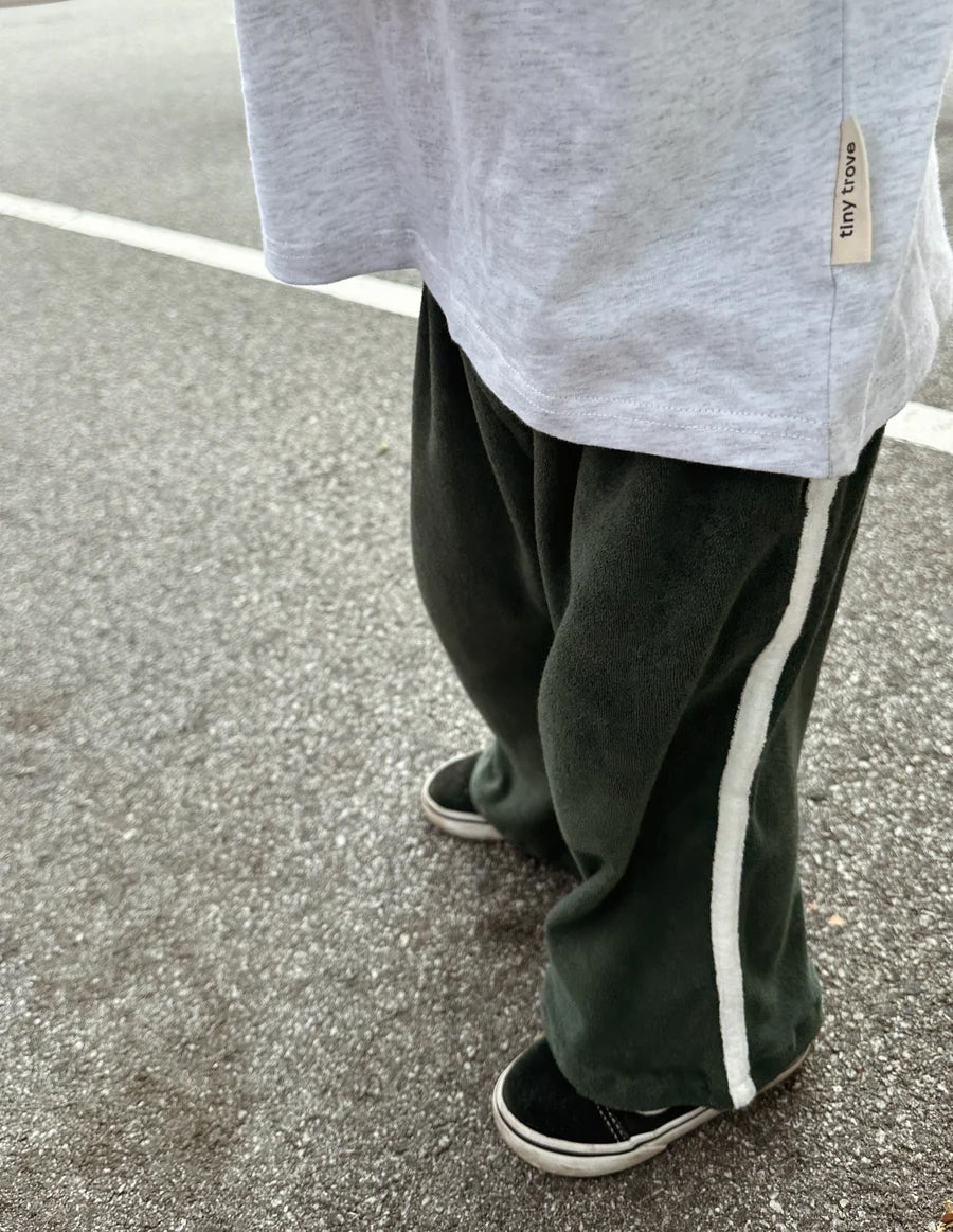 A person wearing TINY TROVE's Darcy Racer Pants in Moss, featuring a dark green color, wide legs, an elastic waistband, and white side stripes, paired with black sneakers stands on a paved surface. The bottom part of the torso is covered by a loose-fitting gray shirt. The ground is gray with a white line running across it.