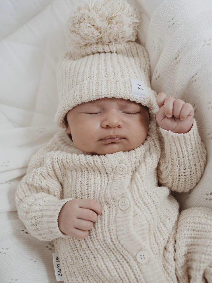 A sleeping baby dressed in ZIGGY LOU's Classic Knit Romper Honey and a matching hat with a large pom-pom is lying on a soft, textured white blanket. The baby has one arm raised, resting on their side, perfectly cozy in their winter outfit.