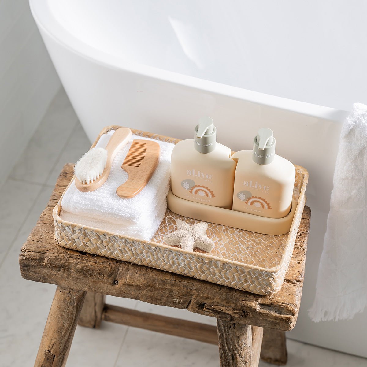 A rustic wooden stool supports a woven basket containing two AL.IVE BABY bottles, a Baby Konjac Sponge - White, a white towel, and starfish beside a cozy bathroom's white bathtub. Ideal for infusing biodegradable elegance into baby bath time.