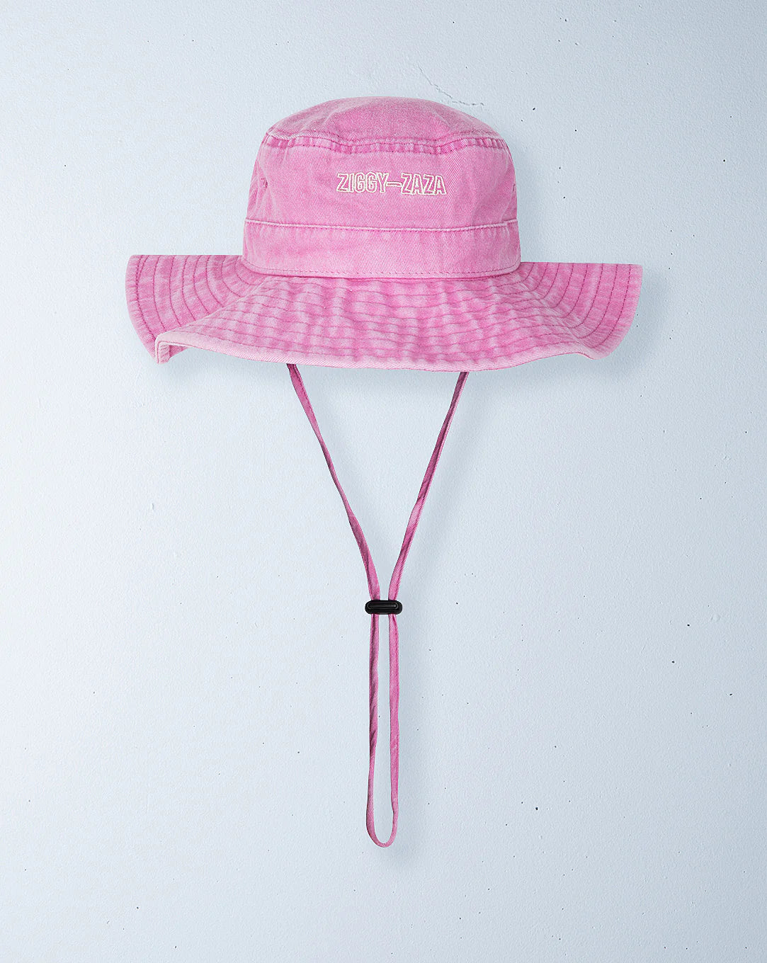 The Ziggy Zaza Everyday Scout Hat Flamingo, a pink cotton canvas hat with a wide brim and adjustable chin strap, featuring embroidered "ZIGGY ZAZA" text, hangs on a light gray wall.