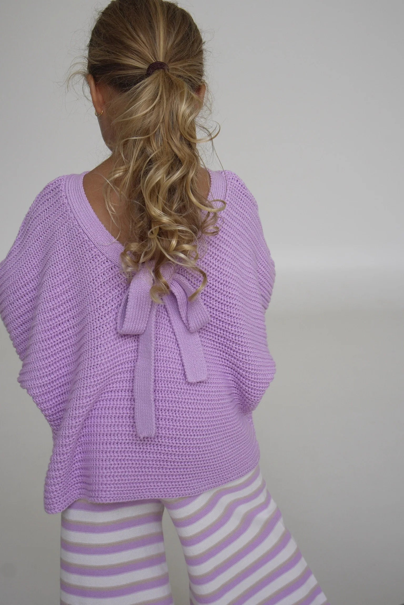 A child with long, curly hair faces away from the camera, wearing a lavender knit top featuring a bow at the back and EN. THE LABEL's Stripe Knit Pants in Lilac, crafted from stretchy fabric. The background is a plain light gray wall.