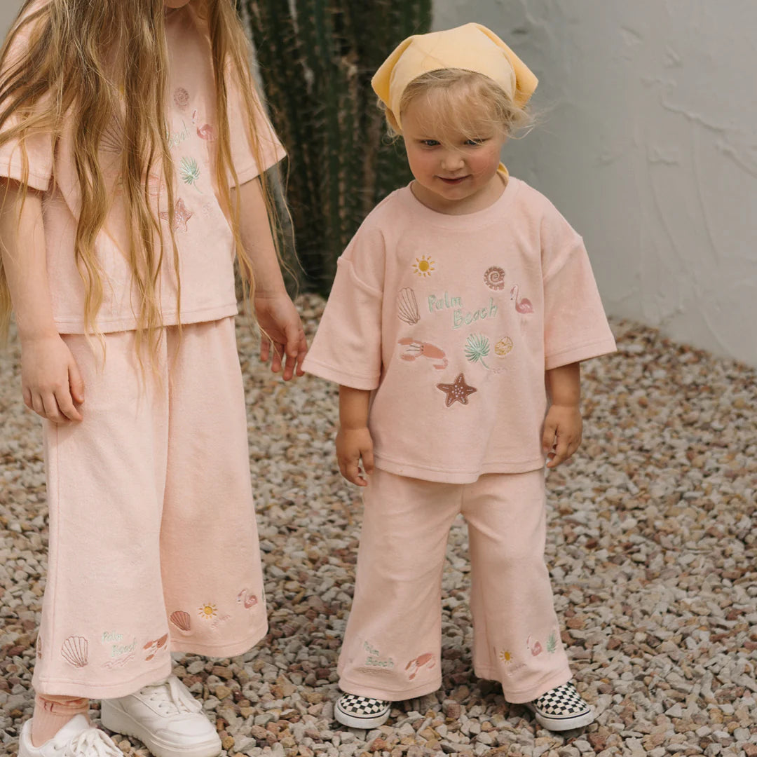 Two young children stand on pebbled ground, wearing matching pastel pink outfits with beach-themed prints. One is dressed in the Palm Beach Wide Leg Pants Terry Towel Palm Flamingo Pink by GOLDEN CHILDREN, paired with a yellow headscarf and checkered shoes. They pose in front of a white wall and a large cactus.