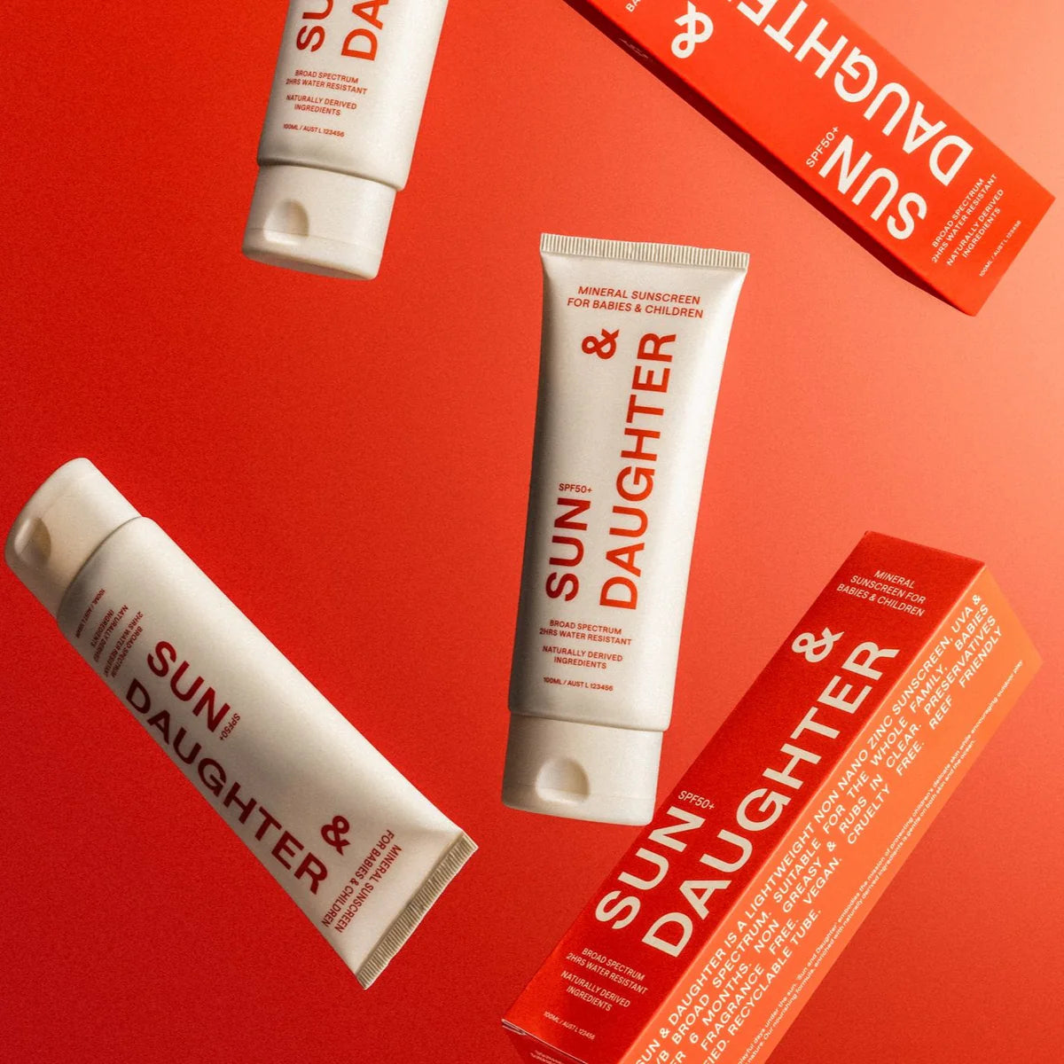 Sun & Daughter SPF50+ Mineral Sunscreen tubes and boxes are set against a vibrant red background. The reef-friendly white and red packaging highlights its suitability for babies and children with SPF 50+ protection.