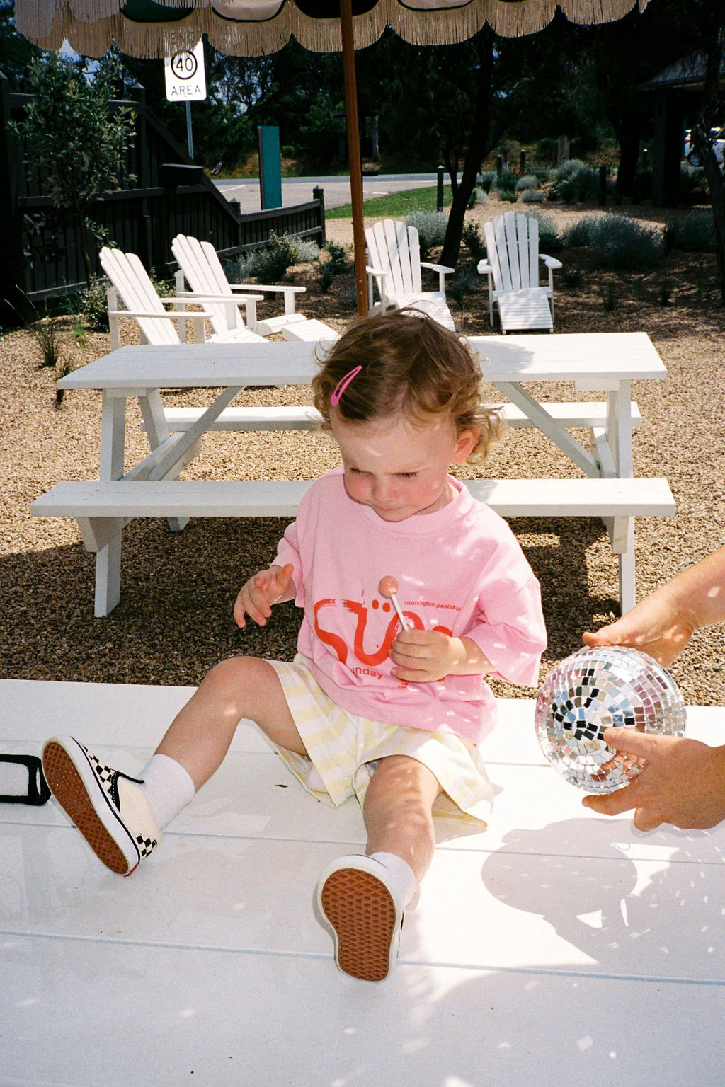 A young child in an oversized Sunday Siblings ~ Il Sole Tee Pink/Red and checkered shoes sits on a picnic table outdoors, holding a lollipop. Nearby, a hand holds a small disco ball. White chairs and an umbrella are in the background, surrounded by gravel and plants.