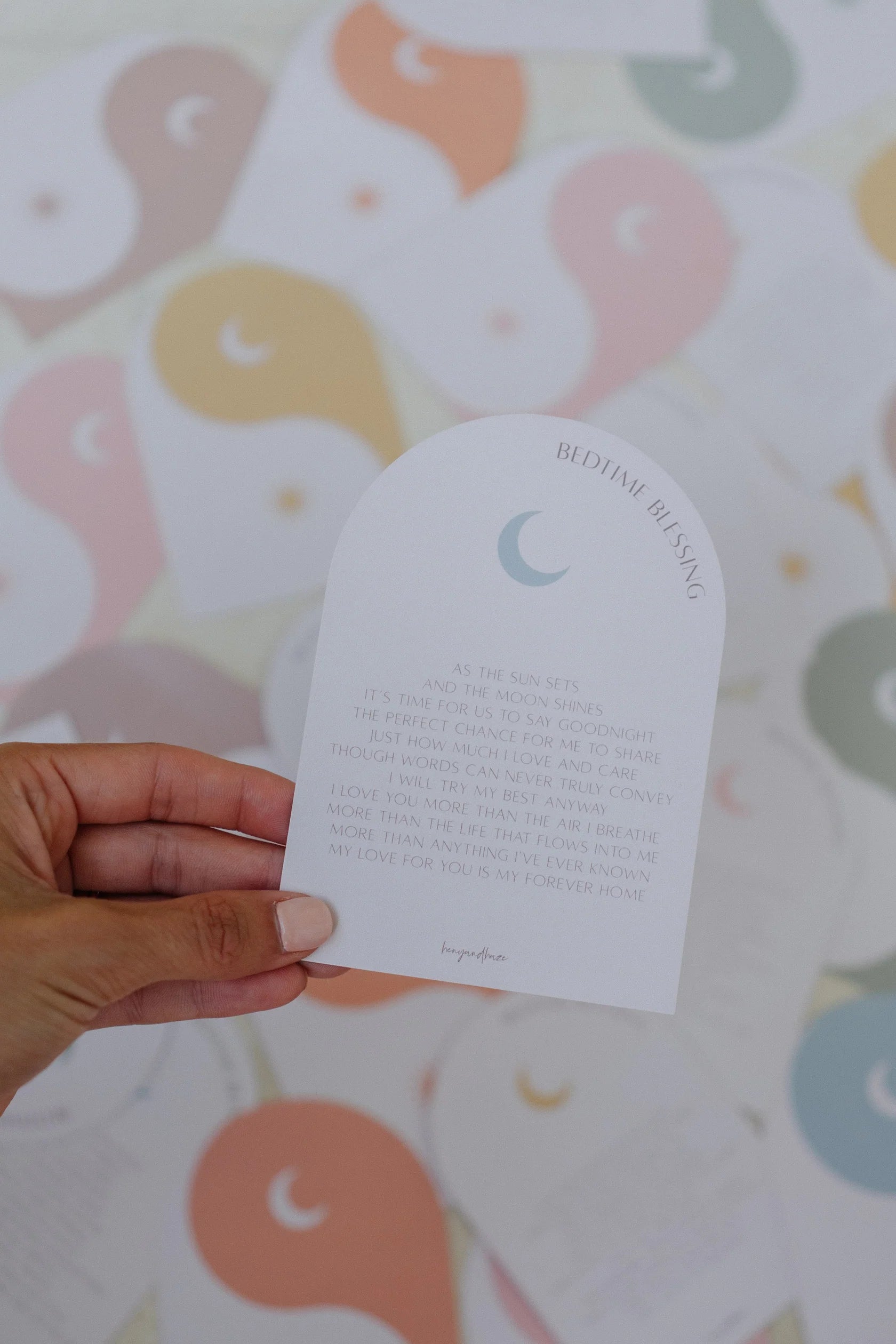 A hand holds a card titled "Bedtime Blessings" from HENY AND HAZE, displaying its text. The blurred background showcases multiple similar cards adorned with pastel yin-yang designs featuring crescent moons. These cards, ideal for tackling kids' sleep challenges, are part of the Bedtime Blessings Deck.