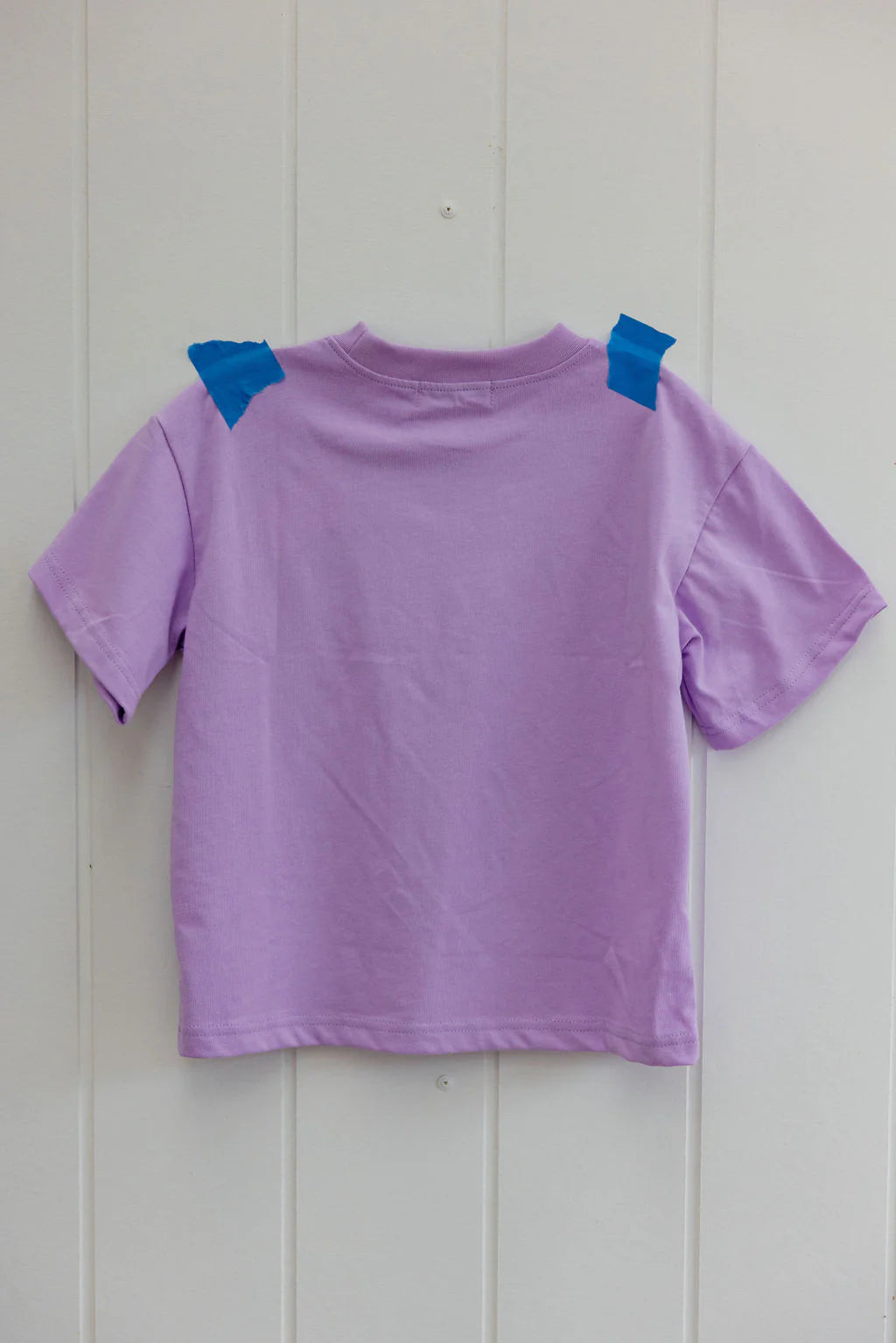 A Sunday Siblings ~ Il Sole Tee in purple/cream, crafted from soft cotton, is taped to a white wall with two pieces of blue tape at the shoulders, displaying the oversized back.