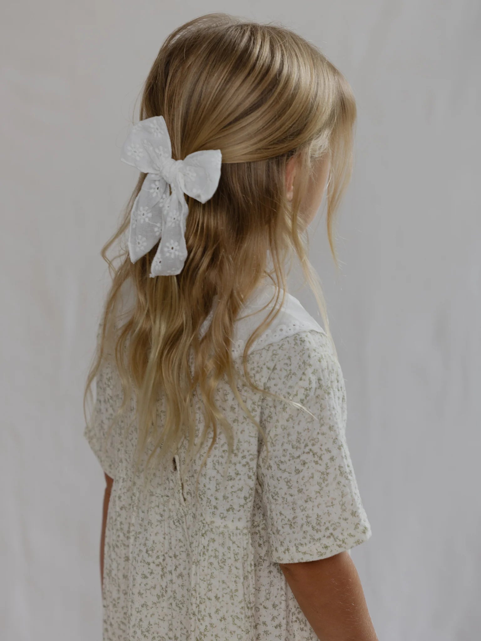 A young girl with long, wavy blonde hair is seen from behind, adorned with the Cotton Bow White hair accessory by VALENCIA BYRON BAY. She is dressed in a light-colored floral dress with a peter pan collar. The background is plain and neutral.