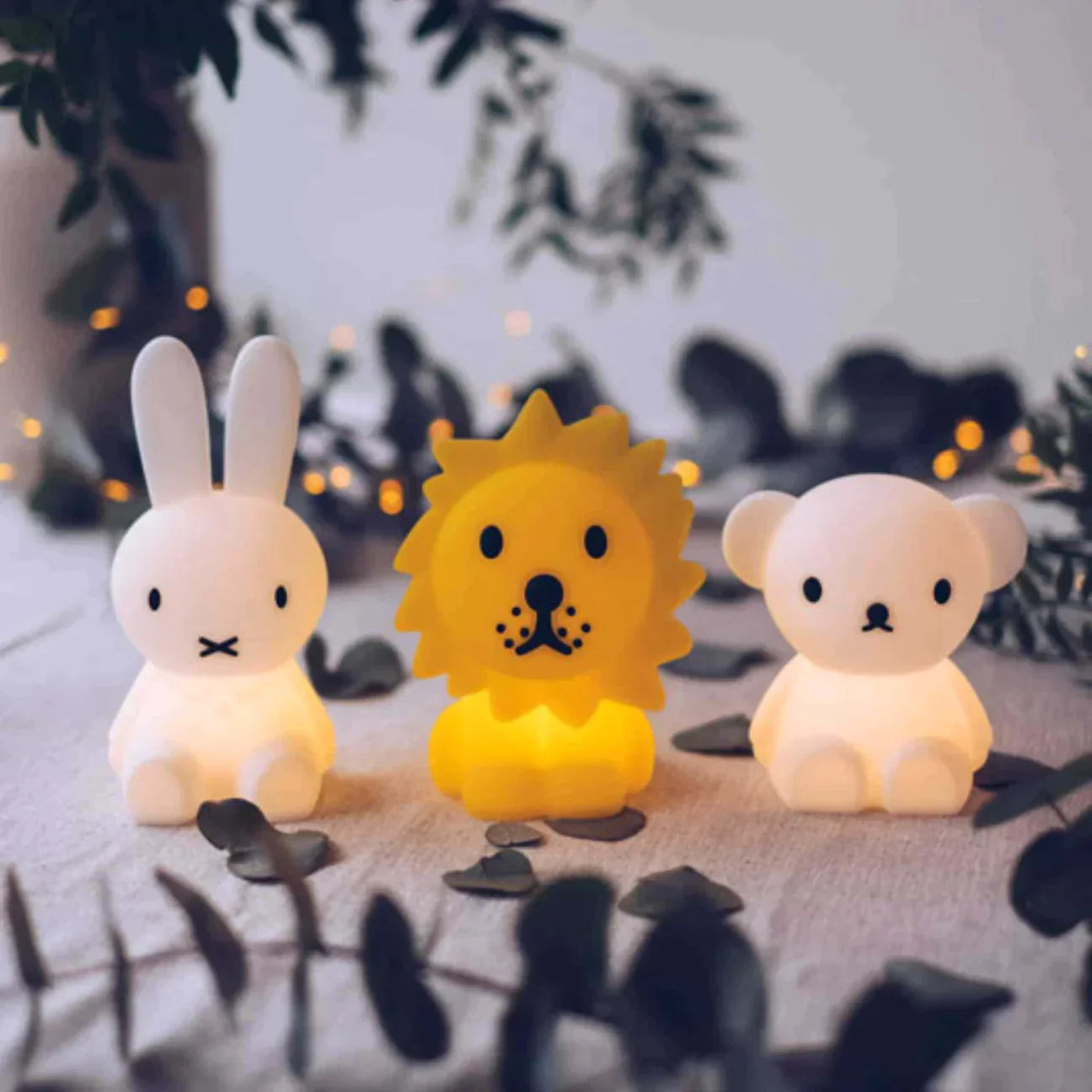 Three adorable animal nightlights are showcased on a table adorned with greenery and delicate fairy lights in the background. From left to right, the soft silicone LED lamps include a white rabbit (MR MARIA's Bundle of Light - Miffy), a yellow lion, and a white bear. The warm glow creates a cozy and charming atmosphere, making these mini-lights perfect collectables.