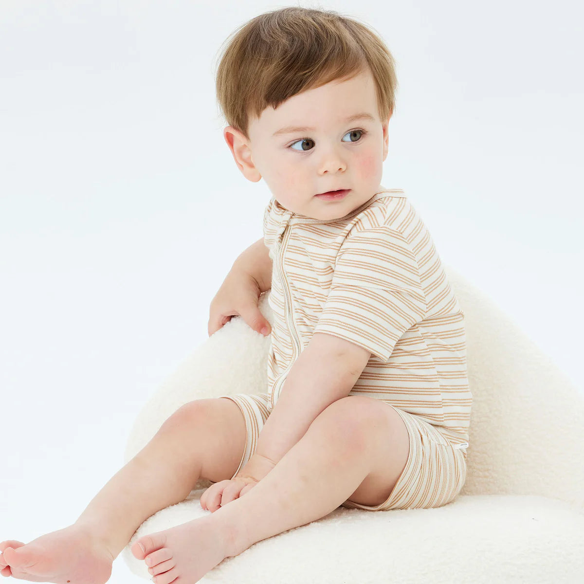 A baby wearing the KYND BABY Day or Night Short Sleeve Onesie Neutral, which is crafted from sustainable viscose fabric, sits on a soft white chair. The baby has light brown hair and is gazing to the side with a curious expression in a plain, white setting.