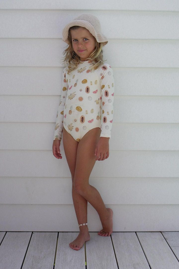 A young girl stands against a white paneled wall, wearing the Tutti Frutti Long Sleeve One Piece swimsuit from EN. THE LABEL, adorned with colorful patterns and paired with a light beige hat. The UFP 50+ rating and chlorine-resistant fabric ensure she stays protected. She has an anklet on one leg and poses with one leg crossed over the other, looking at the camera with a slight smile.