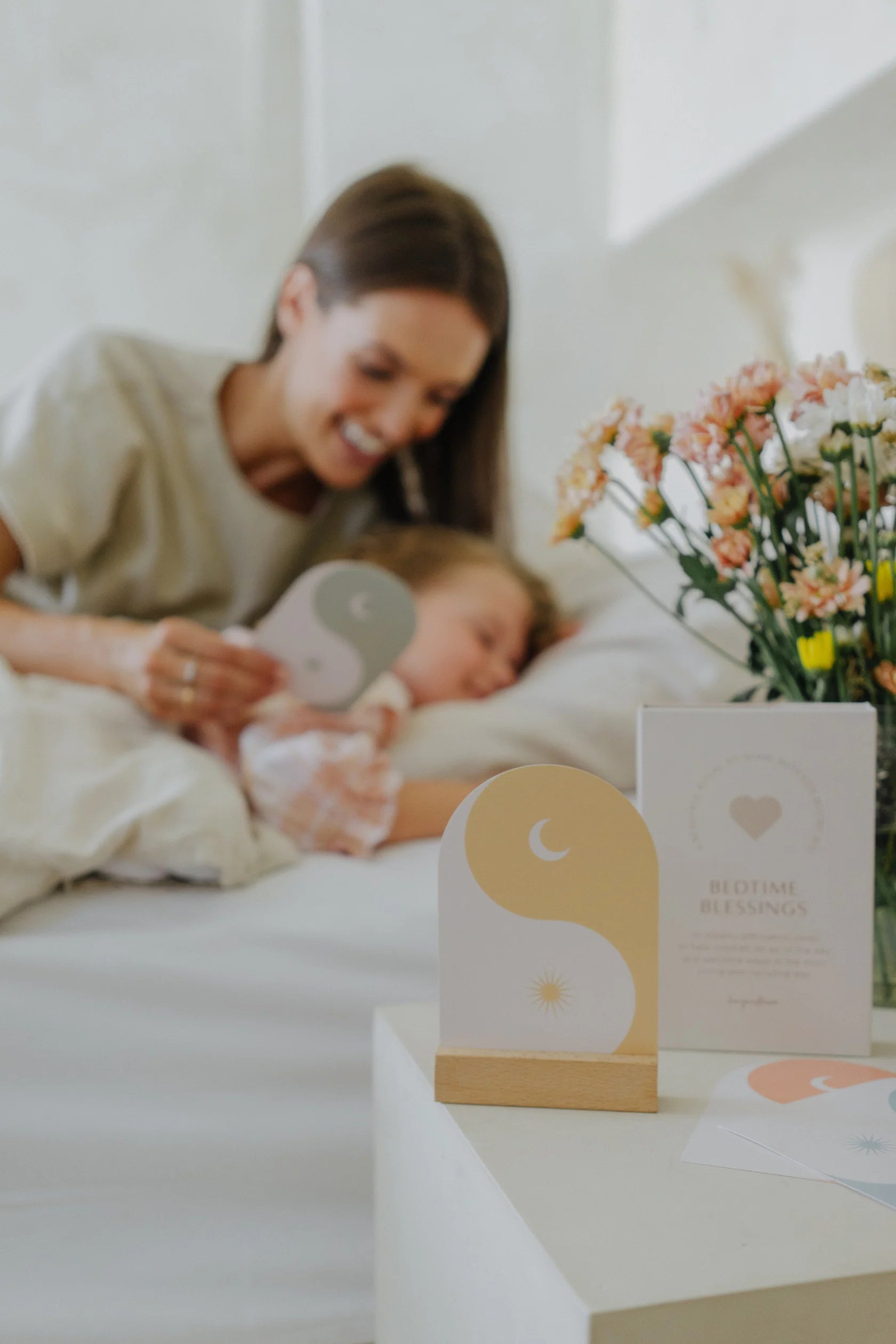 A smiling woman holds a card from the "Bedtime Blessings" deck by HENY AND HAZE while a child lies in bed, seemingly asleep. A bouquet of flowers is on a nearby table, accompanied by another card that reads "Bedtime Blessings," adorned with a heart design. The scene exudes calm and serenity, suggesting an affirming bedtime routine.