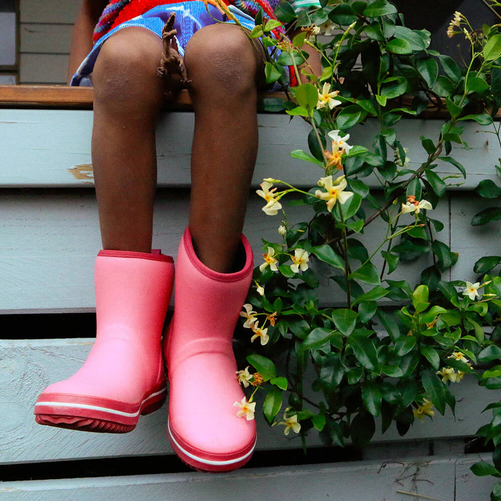 A child in Merry People ~ Andie Kids Gumboot Bubblegum boots sits on a step beside a leafy plant with yellow flowers. These vibrant pink, waterproof, vegan-friendly boots ensure they stay dry and eco-conscious, even on rainy days.