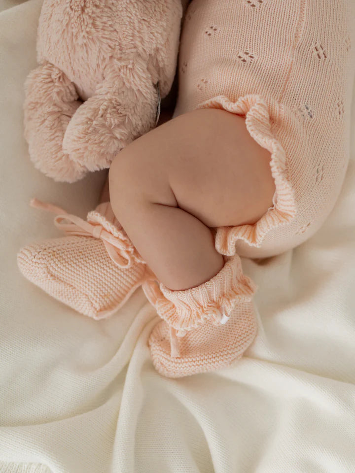 A baby dressed in a ZIGGY LOU Frill Bodysuit Primrose with matching booties is lying on a cream-colored blanket. The baby's legs are curled up, and a soft, light pink stuffed animal is partially visible beside the baby.