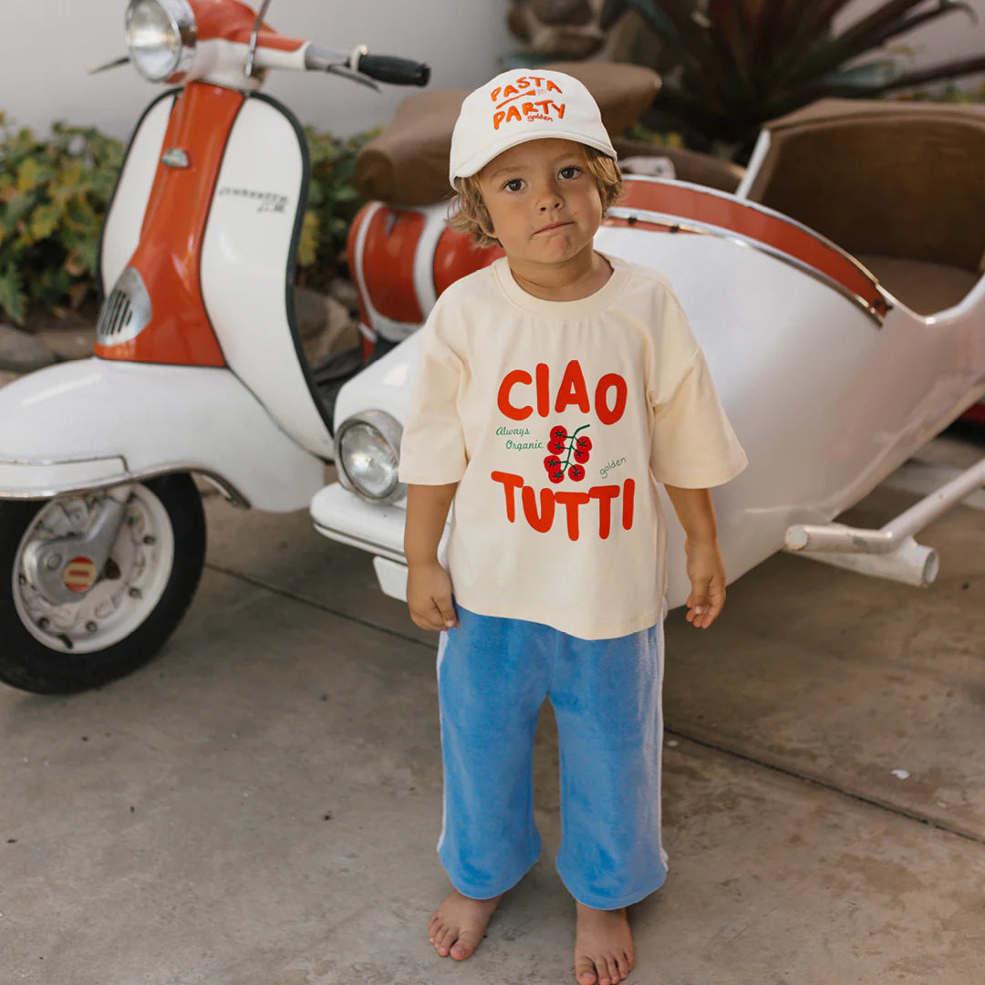 A young child in a "CIAO TUTTI" shirt and "PASTA PARTY" hat stands barefoot beside a vintage red and white scooter with a sidecar, wearing Golden Children’s Ciao Tutti Relaxed Pants Terry Towel Pool Blue. Lush plants enhance the outdoor setting.