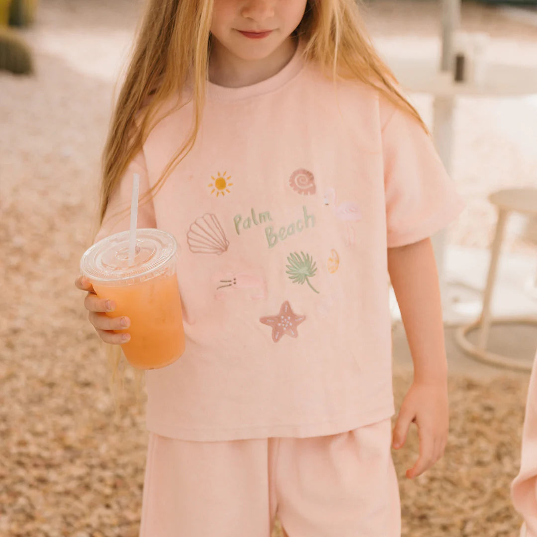 A young girl in a Flamingo Pink Palm Beach Mid Sleeve Tee by GOLDEN CHILDREN, featuring beach-themed illustrations and subtle embroidery details, holds a plastic cup of orange drink. She stands on a pebble-covered ground, exuding summertime vibes.