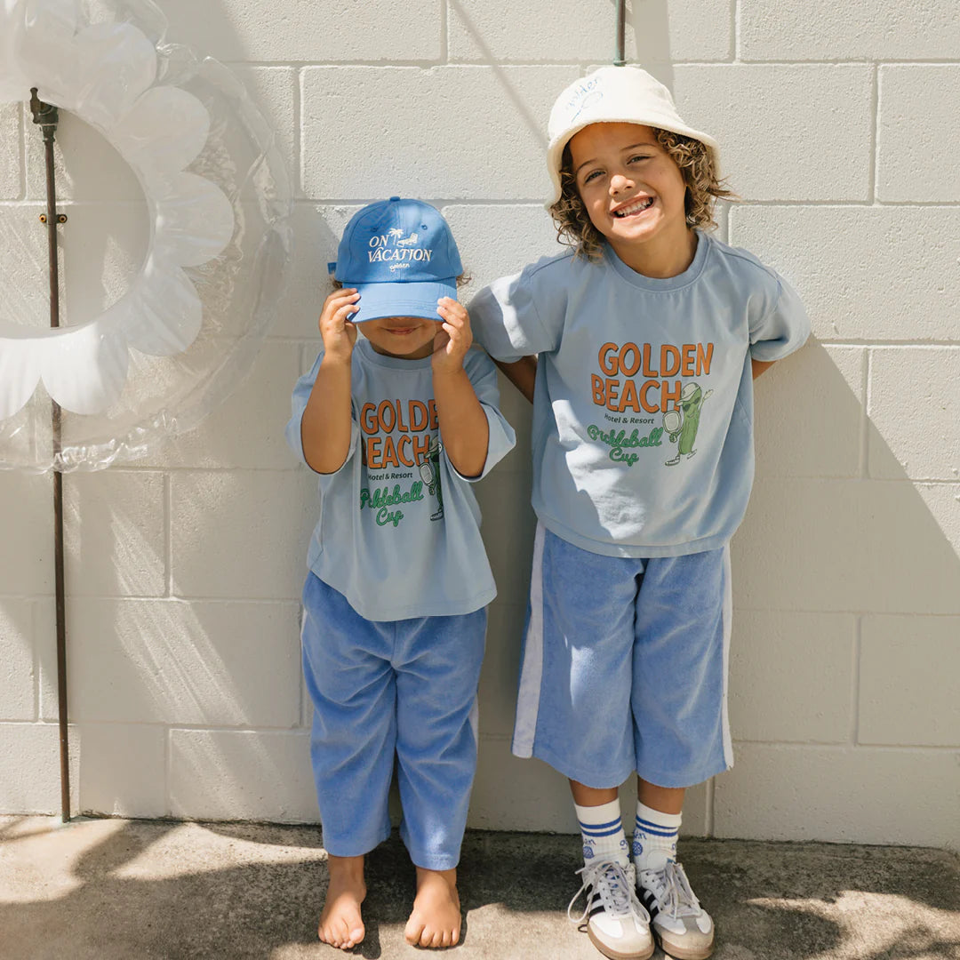 Two children wearing matching Golden Beach Mid Sleeve Tee Palm Blue shirts by GOLDEN CHILDREN stand against a white brick wall. Both have hats, and the child on the left is adjusting theirs. The relaxed fit of their outfits perfectly complements the sunny day vibe.