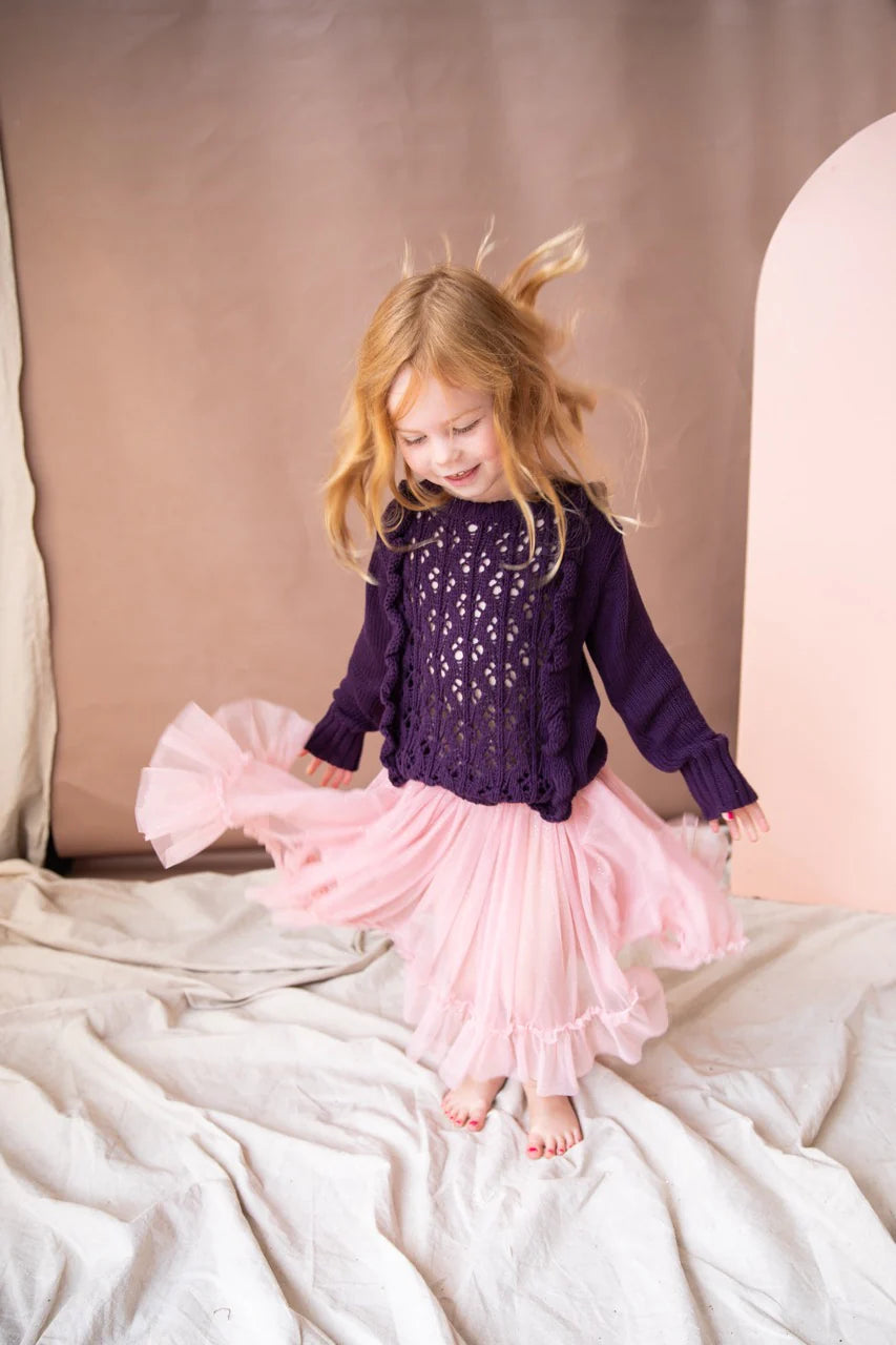 A young girl wearing the carrie tutu in pink lady apple colour. The girl is wearing a purple knitted jumper and twirling around in circles. There are cream drapes on the wall and ground.