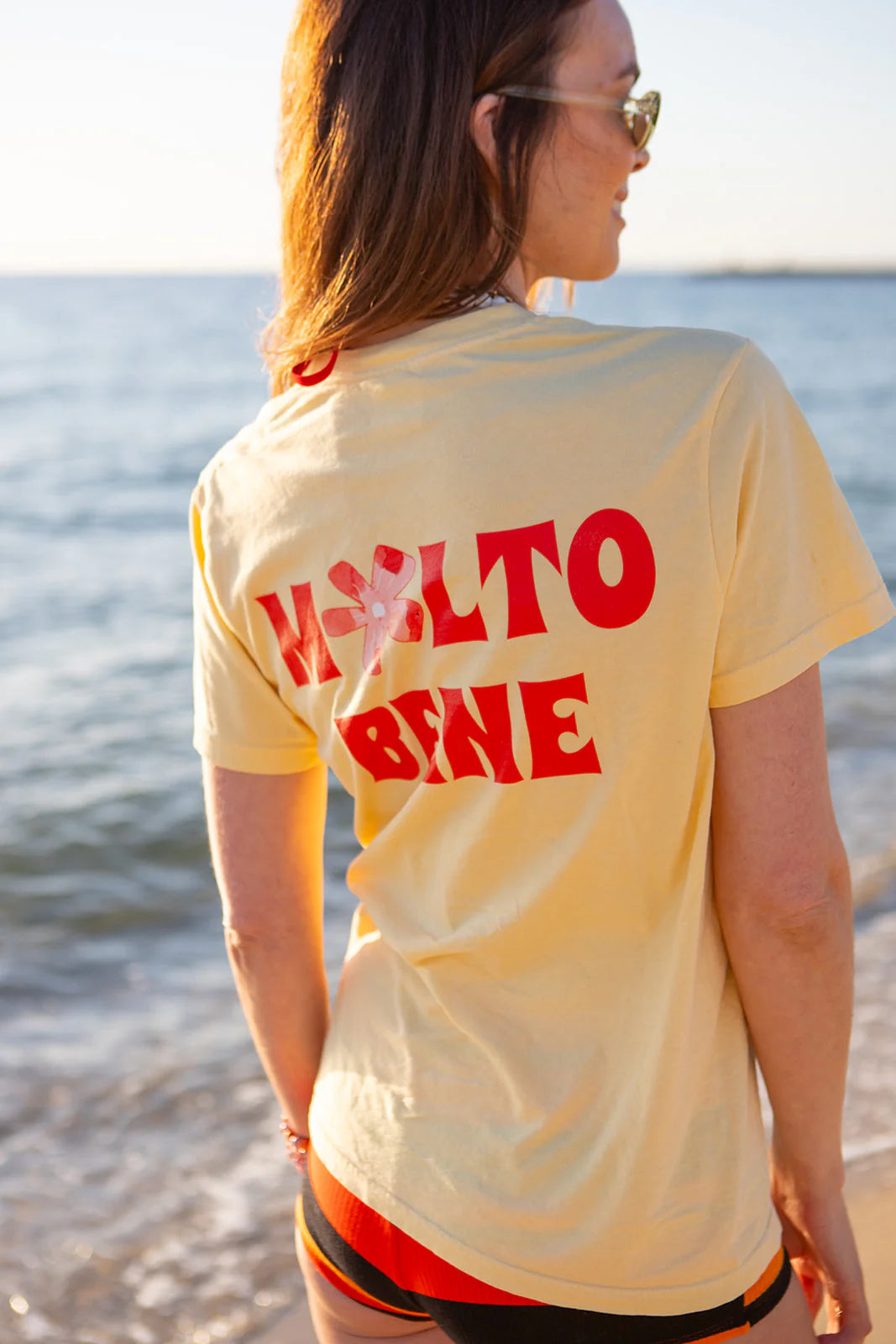 A person with long hair and sunglasses stands on a beach wearing a yellow Molto Bene T-Shirt by MAKU THE LABEL. It features "MOLTO BENE" in red and hand-painted flower designs on the back, made of 100% cotton, illuminated by the shining sun.