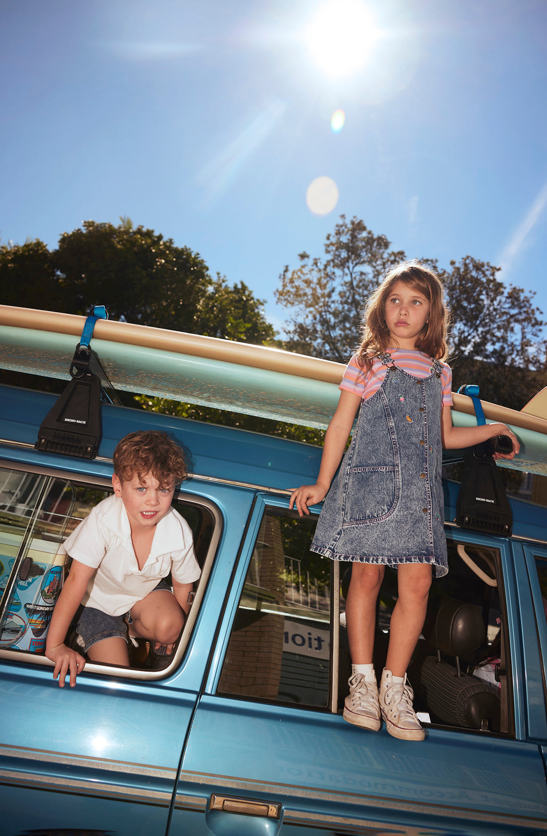 Two children are playing on a vintage blue van. The boy with brown curly hair, dressed in a white shirt and shorts, is climbing out of the van window. The girl with long brown hair, wearing a pink shirt and the Penelope Fruity Denim Pinafore Dress by GOLDIE + ACE with adjustable brass straps, is standing on the van's sideboard. Sunlight shines brightly overhead.