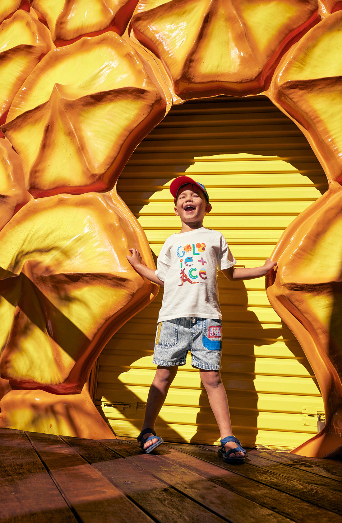 A young child stands joyfully in front of a bright yellow backdrop resembling a pineapple slice. The child wears a red cap, a white t-shirt with colorful text, light denim Noah Colour Pop Denim Shorts from GOLDIE + ACE with an elastic waistband and functional drawcord, and sandals, with hands spread out against the pineapple background.