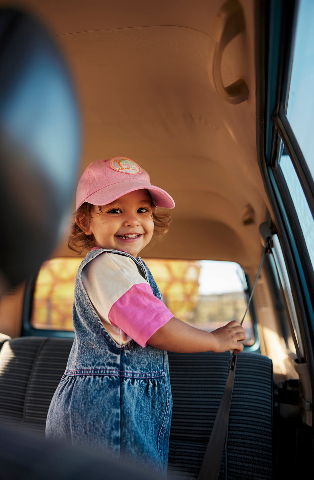 A smiling young girl wearing a pink baseball cap, a pink and white shirt, and the Penelope Fruity Denim Pinafore Dress by GOLDIE + ACE stands inside a car. She is holding onto the seatbelt with one hand and looking directly at the camera while sunlight illuminates the car's interior.