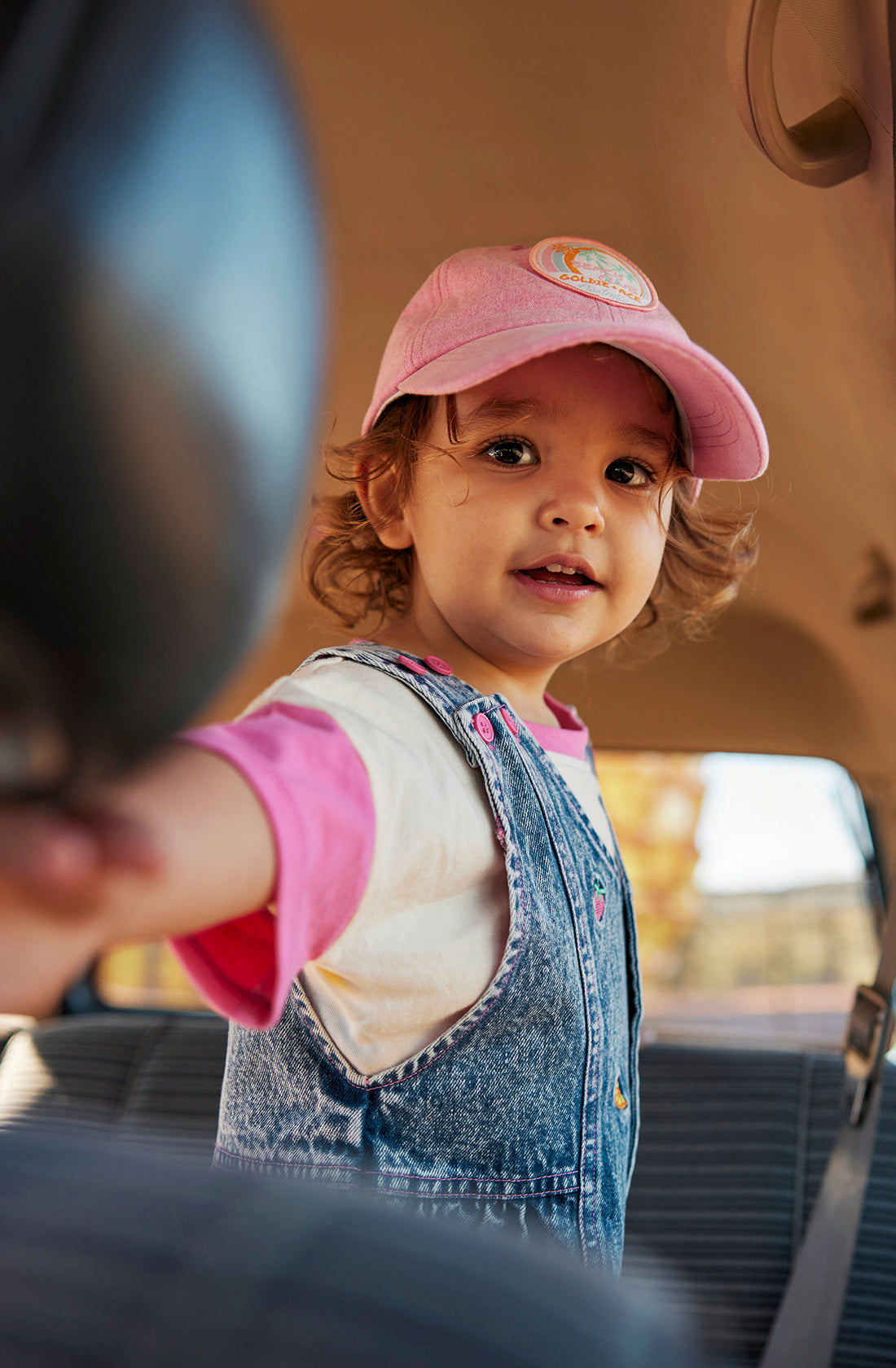 A toddler wearing the Penelope Fruity Denim Pinafore Dress by GOLDIE + ACE smiles while standing inside a vehicle. The child reaches towards the camera with one hand, and the background through the windows shows a sunny outdoor scene.