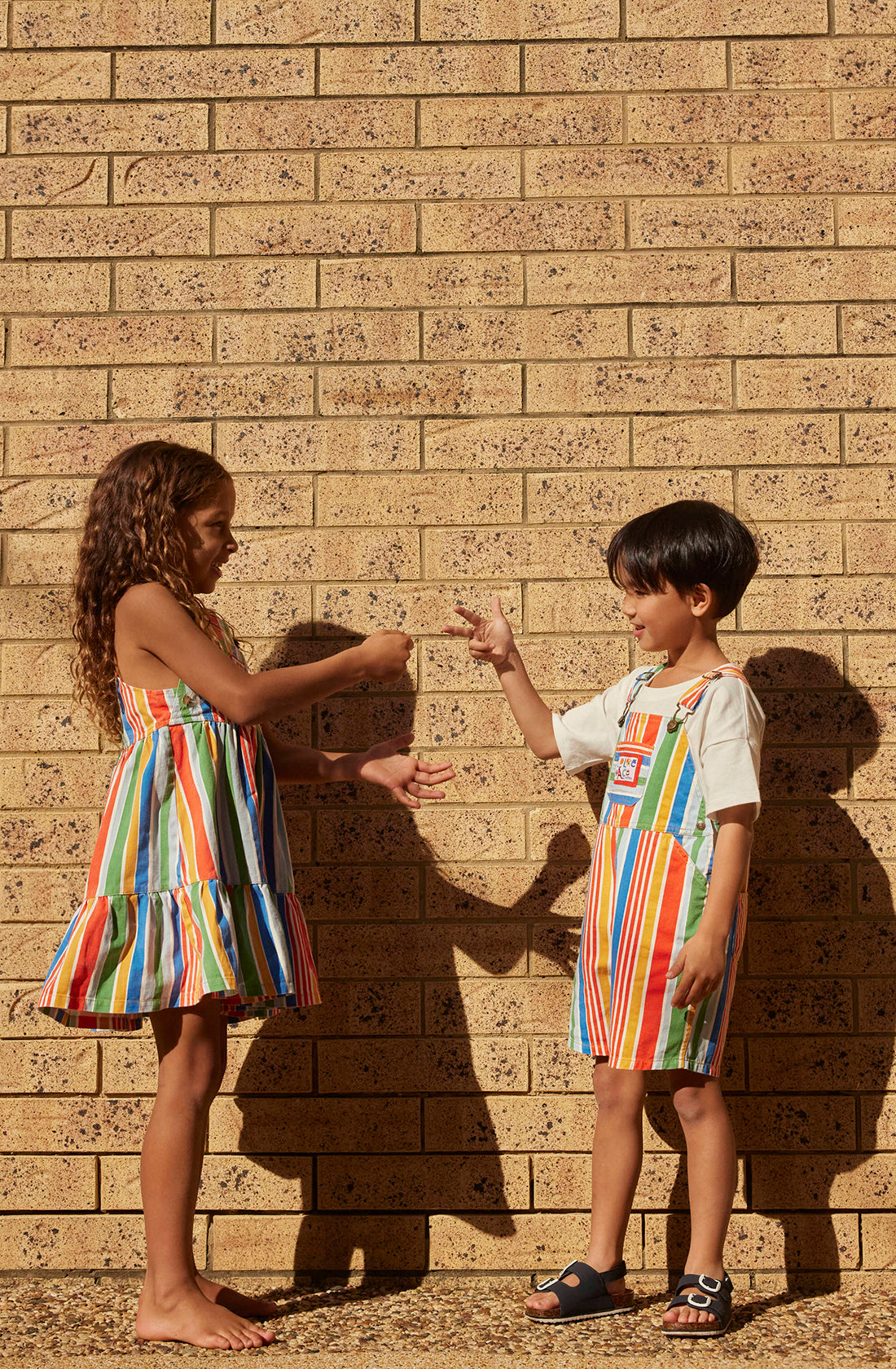 Two children, a young girl and boy, stand against a brick wall on a sunny day. The girl wears the Tully Tiered Pinafore Dress from GOLDIE + ACE, a colorful striped outfit with adjustable straps and brass snap buttons. Both are engaged in playing rock-paper-scissors, smiling and looking at each other's hands. Their shadows are cast on the wall behind them.