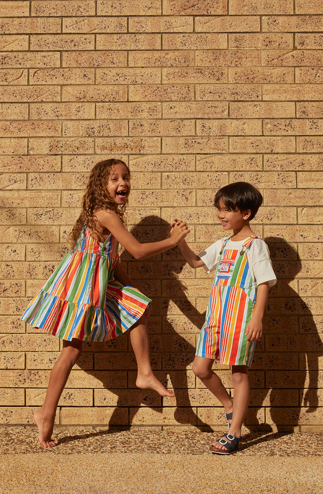 Two children, a boy and a girl, joyfully high-fiving in front of a brick wall. The girl wears the Tully Tiered Pinafore Dress with colorful stripes by GOLDIE + ACE, while the boy is in blue denim overalls with brass snap buttons and adjustable straps. Sunlight casts their shadows on the wall.