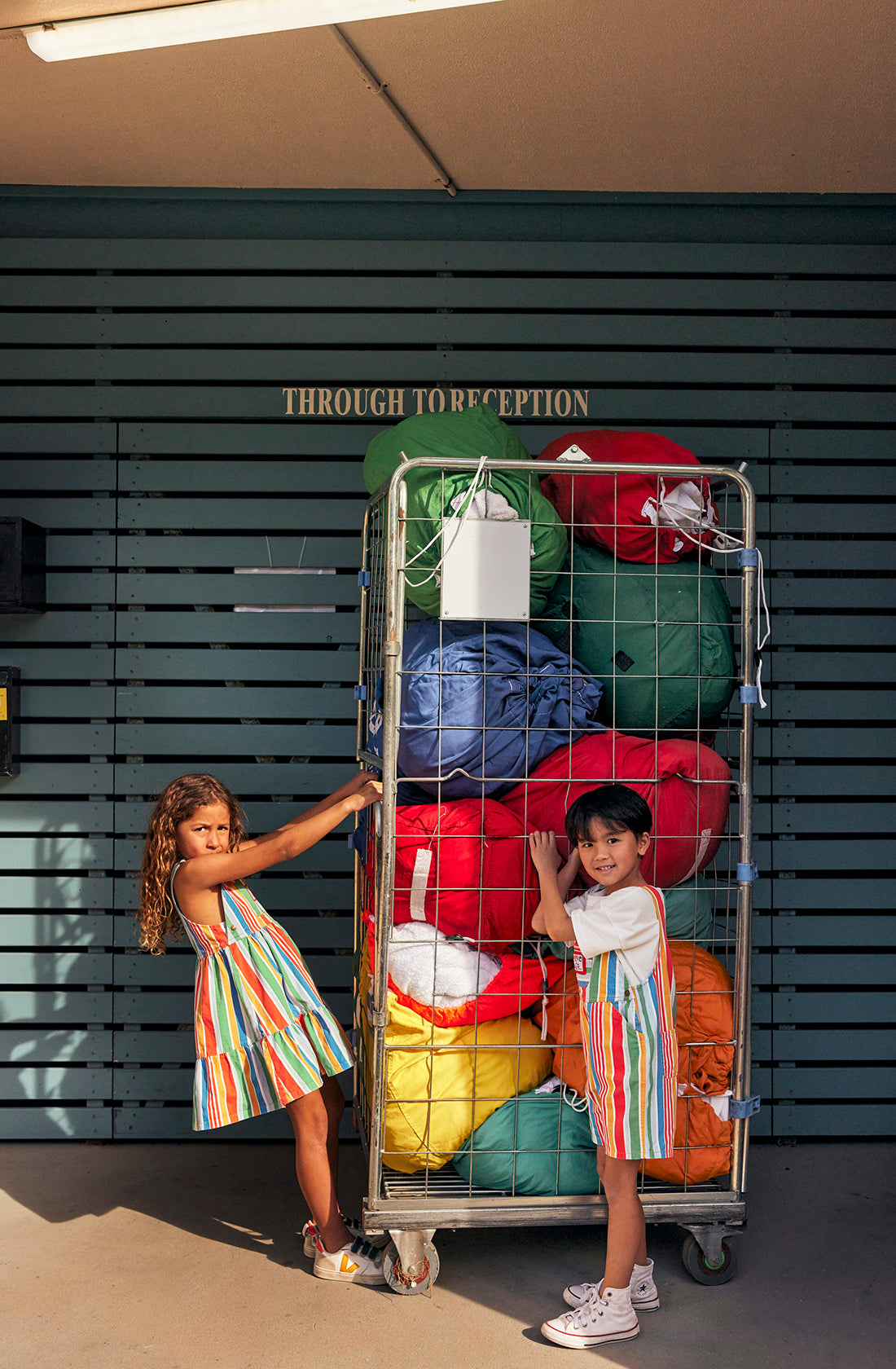 Two children wearing matching Burton Striped Denim Overalls by GOLDIE + ACE stand next to a large metal cart filled with rolled sleeping bags in various bright colors. They are smiling and posing playfully in front of a building with a sign that reads "Through to Reception.