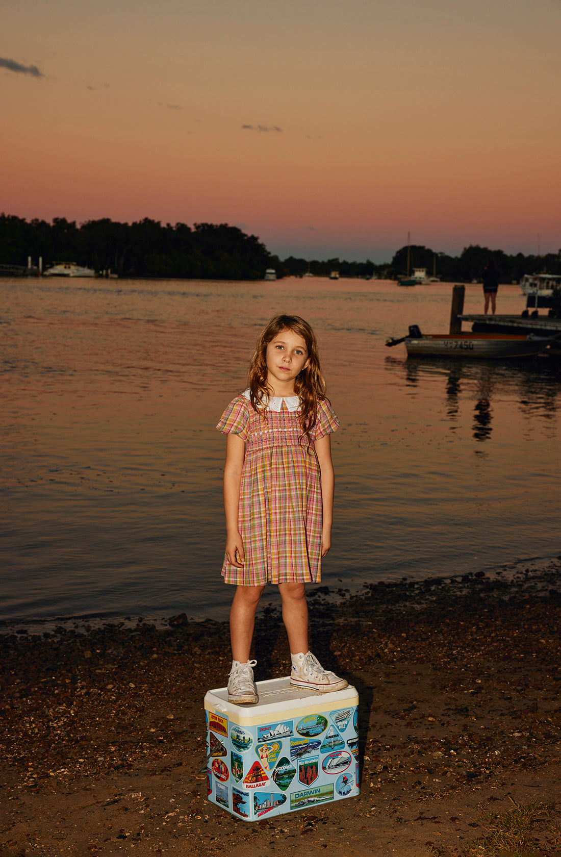 A young girl in the Flo Smocked Dress by GOLDIE + ACE stands on a cooler decorated with stickers near the edge of a calm body of water at sunset. The sky is tinged with hues of pink and orange, and trees and boats are visible in the background. She gazes directly at the camera.