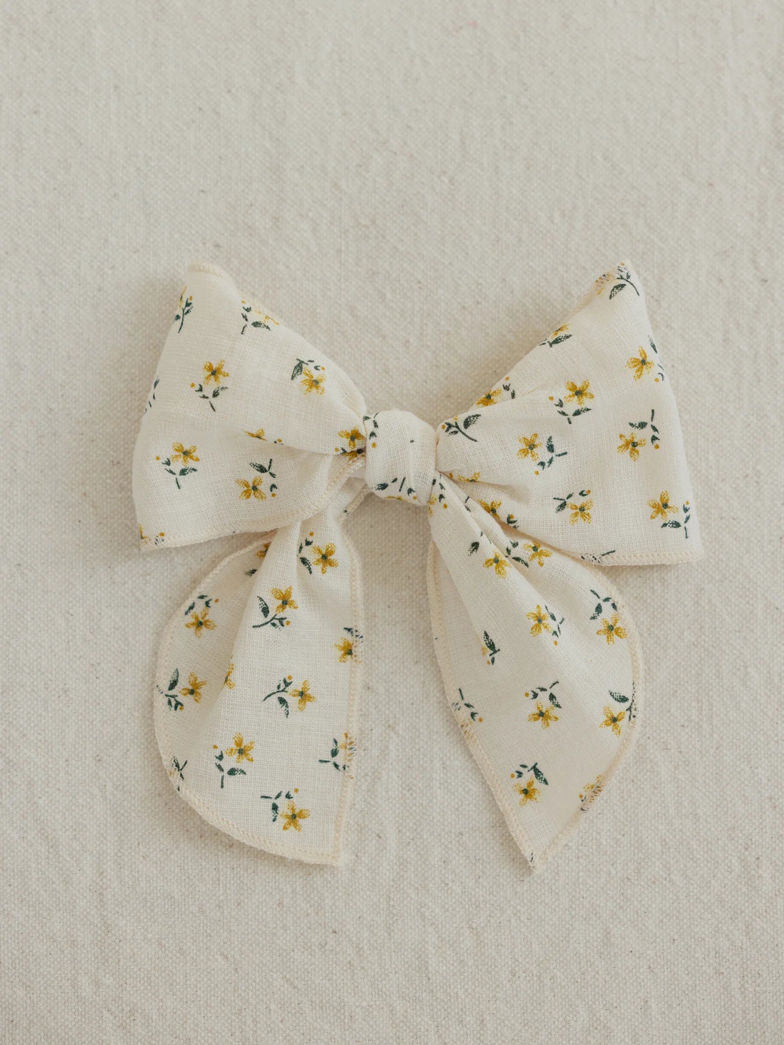 The VALENCIA BYRON BAY Cotton Bow Marigold, handcrafted from cream-colored cotton adorned with small yellow flowers and green leaves, is set against a light beige, textured background—ideal as a charming gift or a stylish hair accessory.