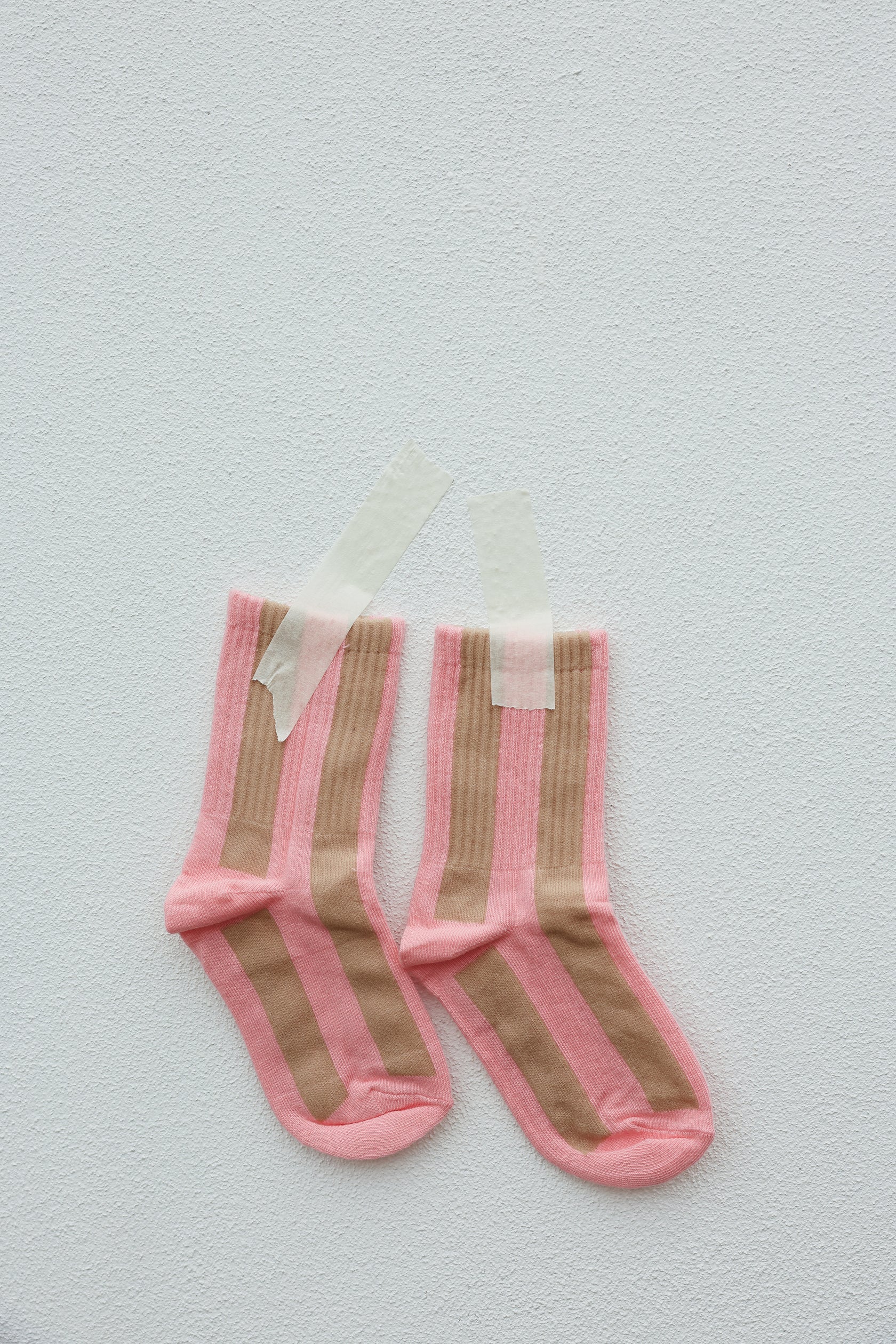 A pair of Circus Sock from TINY LOVE CLUB, designed with pink and beige stripes and ideal for children aged 2-5 years, is taped to a white wall using two pieces of masking tape. Each small sock features a ribbed texture near the top, with the backdrop being a textured white surface. And if you're wondering about maintenance, don't worry—they are machine washable.