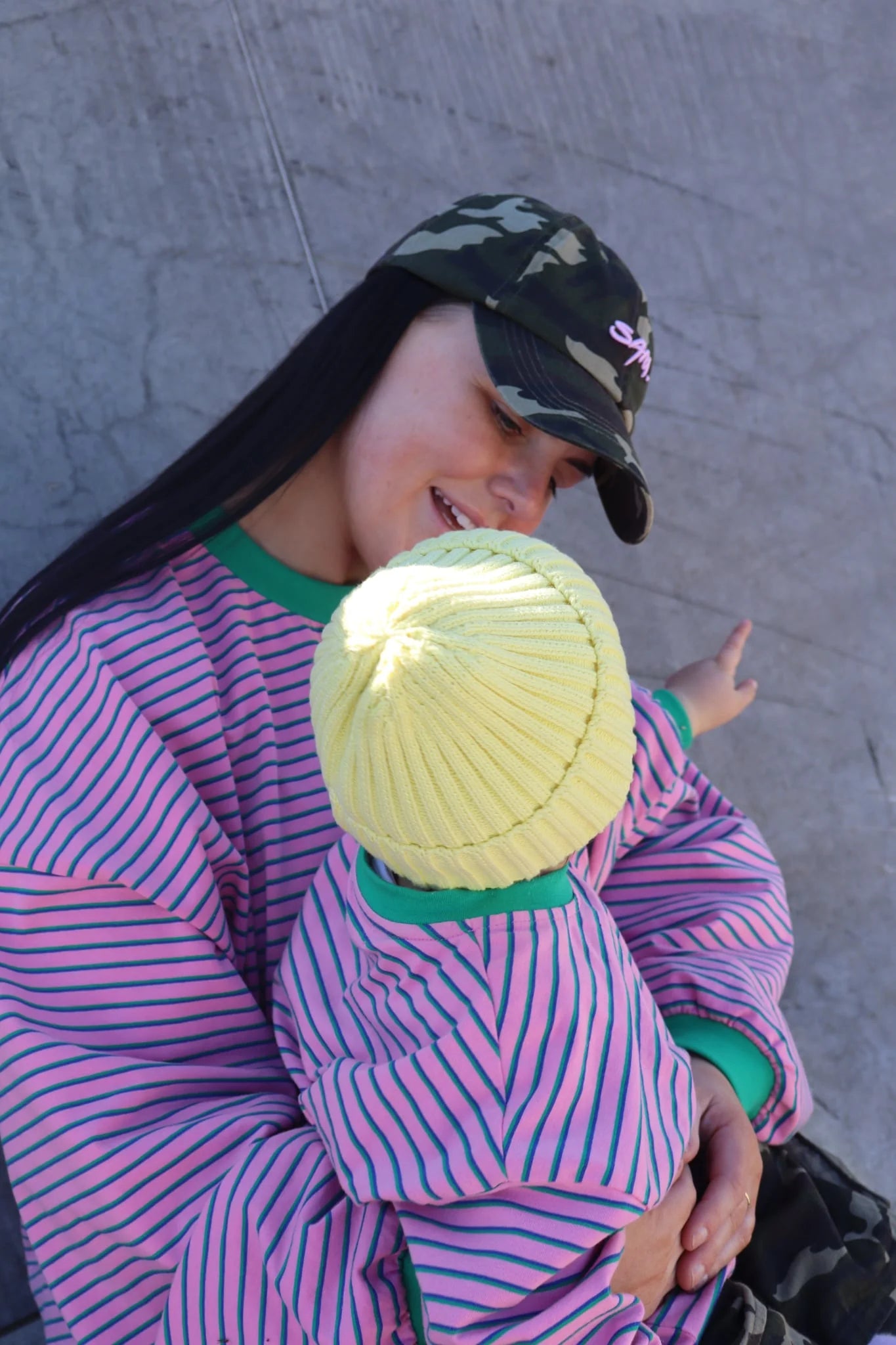 A woman in a striped pink shirt and camo hat holds a small child wearing the Sam the Label - Kids Watermelon Sugar LS Tee by SAM THE LABEL and a yellow knit hat. The child points away as they both smile outdoors.