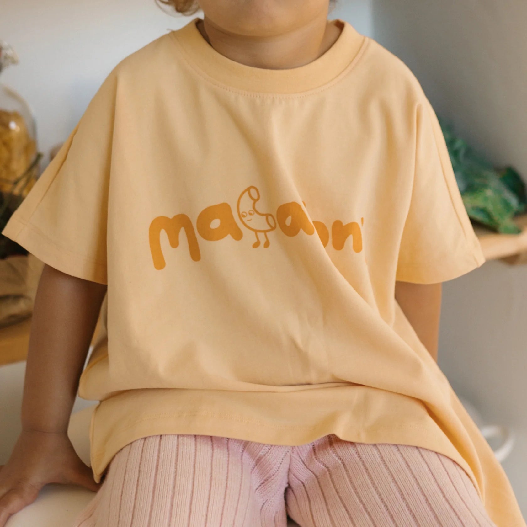 A young child is wearing an oversized, light peach-colored Macaroni Tee Golden Butter from GOLDEN CHILDREN made from soft stretch cotton jersey, featuring an orange graphic and the word "macaroni" in playful letters on the front. The child is seated and also dressed in light pink ribbed pants. The background includes some indistinct objects and greenery.