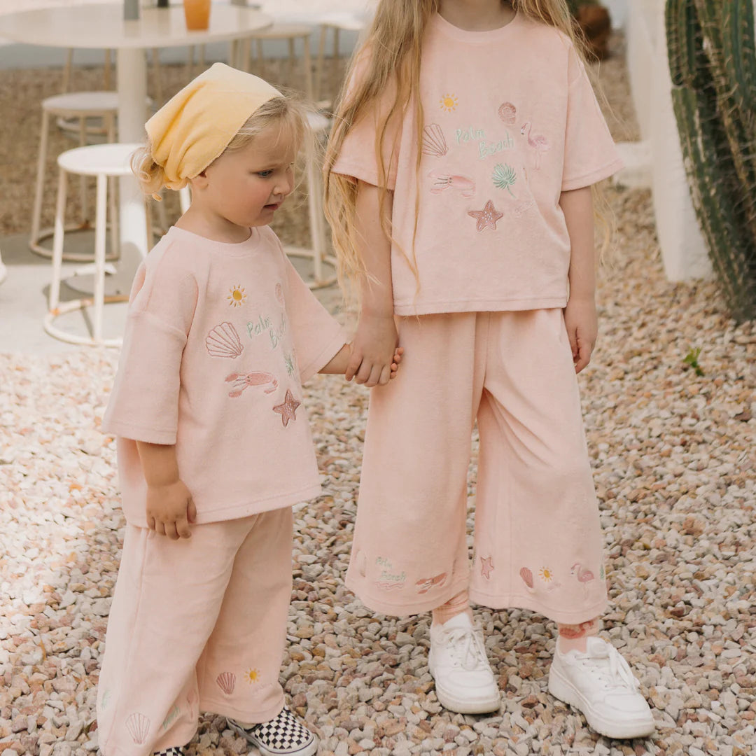 Two children are holding hands, wearing matching GOLDEN CHILDREN Palm Beach Mid Sleeve Tee Terry Towel shirts in Flamingo Pink. They stand on a pebbled ground next to a potted cactus, with one child sporting a yellow headscarf and both wearing light-colored shoes.