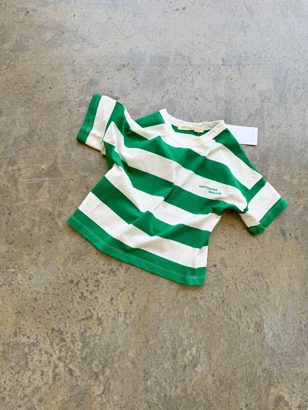 The Twin Collective ~ Stripe Tee Green by TWIN COLLECTIVE, a green and white striped kids' short-sleeve shirt made from 100% cotton, sits on a concrete surface. It has a small logo near the chest and a tag on the neckline.