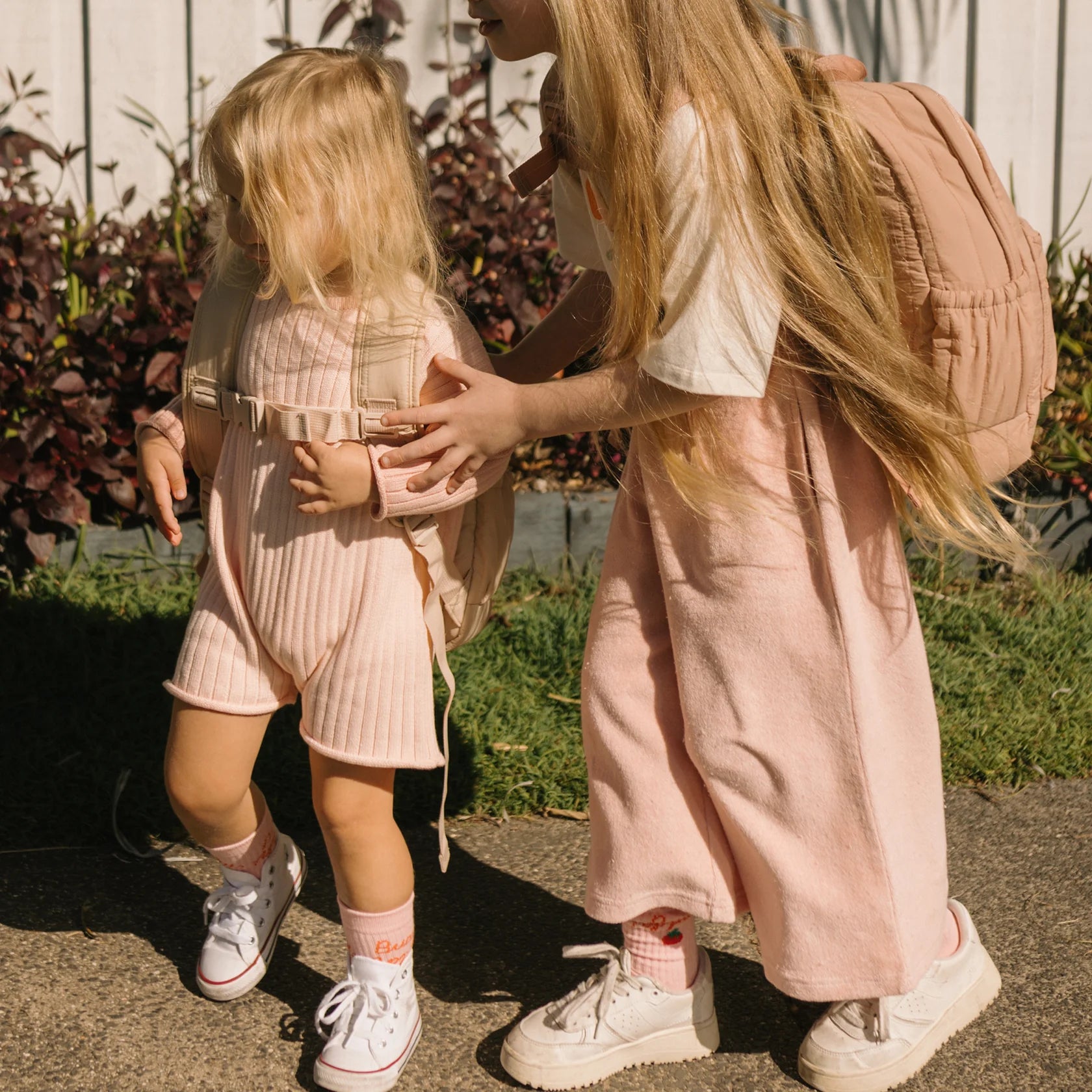 Two children with long blonde hair, dressed in GOLDEN CHILDREN Confetti Wide Leg Pants Pink Parfait and white sneakers, stand outdoors. The older child, wearing a dress and a backpack, gently helps the younger child in a romper and backpack. They stand on a path next to plants.