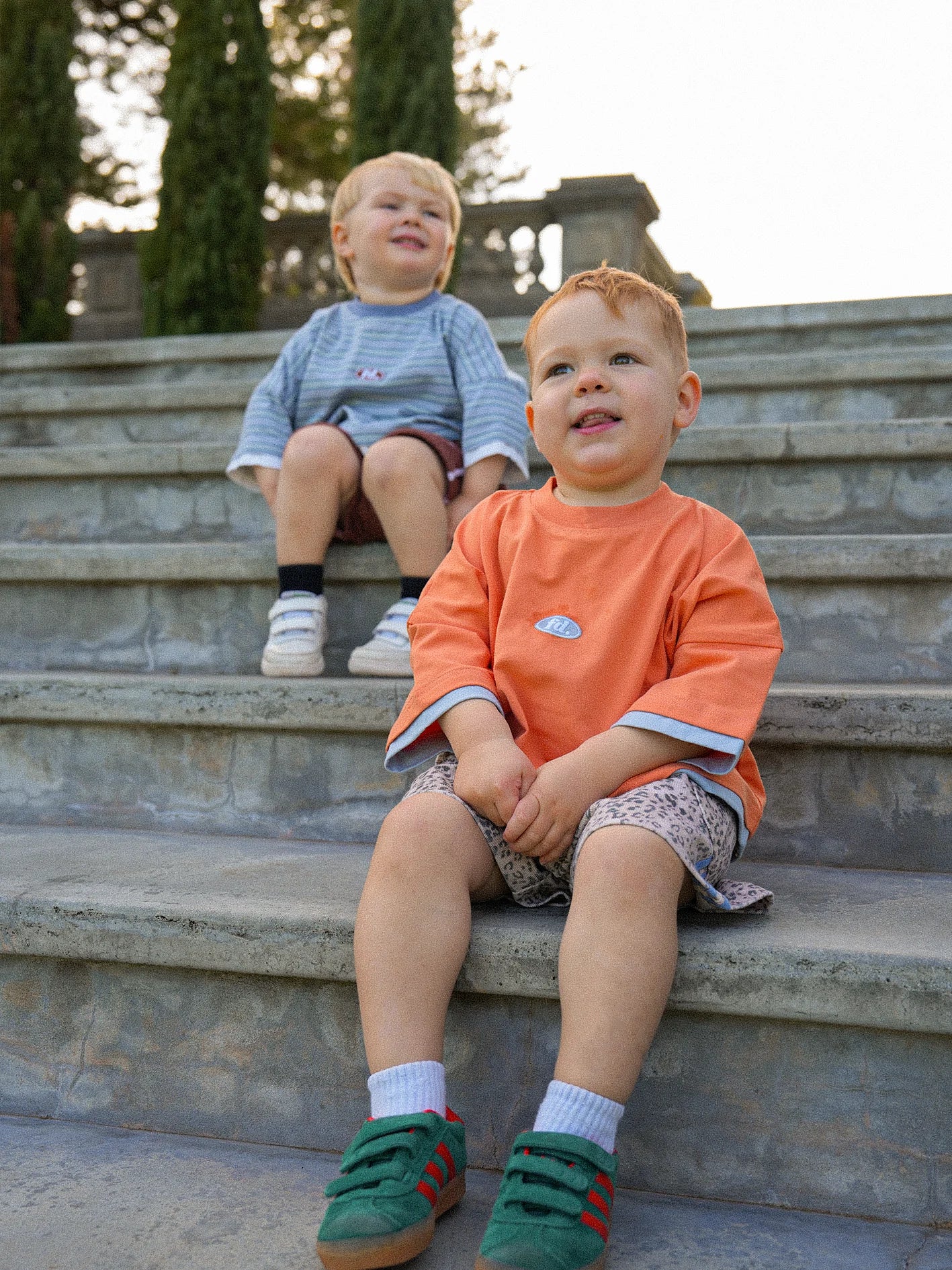 Two toddlers sit on stone steps outdoors, dressed in casual attire. One is wearing a PRE-ORDER Double Layer Tee Paw Paw by FRANCO'S DAD paired with patterned shorts, while the other sports a striped shirt and navy shorts. Both are smiling, surrounded by trees and a stone railing in the background.