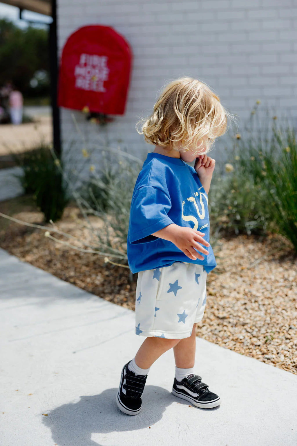 A young blond child in an oversized SUNDAY SIBLINGS Il Sole Tee Blue/Yellow and star-patterned white shorts walks on a sidewalk, looking down with one hand near their mouth. Greenery surrounds them, and a blurred red sign is in the background.