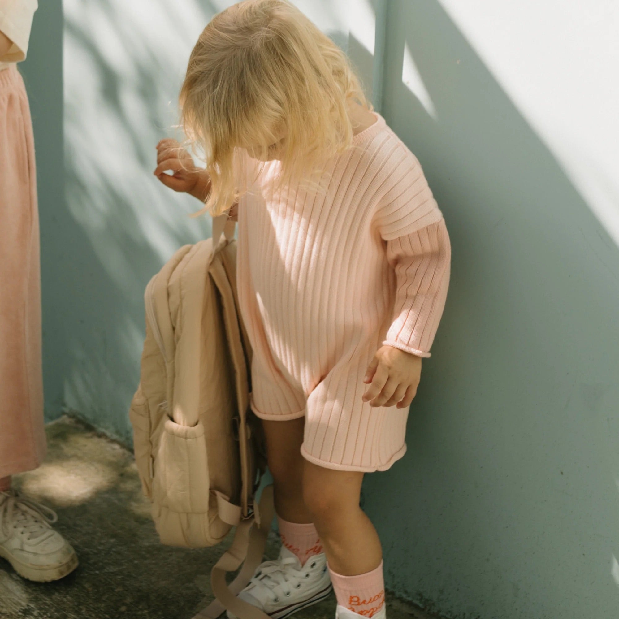 A toddler with blond hair, wearing a Golden Knit Romper Strawberry Sundae from GOLDEN CHILDREN and matching socks, looks down while holding a beige backpack. The child stands next to an adult wearing light-colored clothing and sneakers. The background features a light blue wall and dappled sunlight.