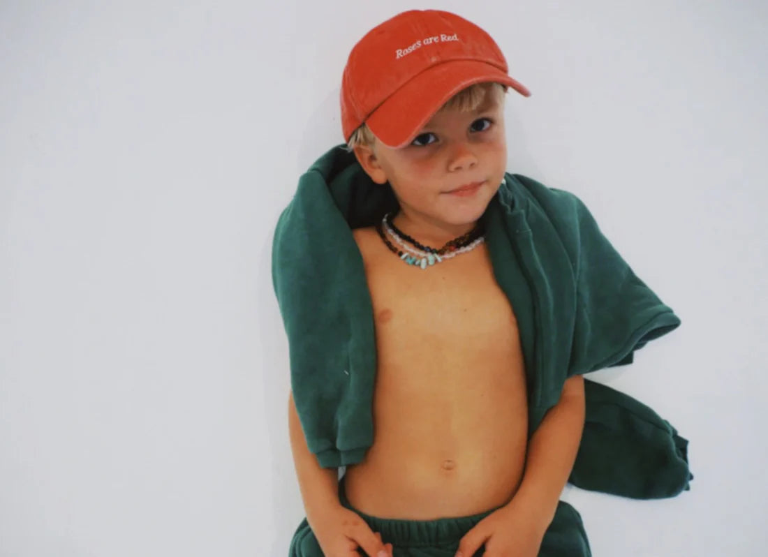 A young child stands against a white background, wearing the stylish Ziggy Zaza ~ Roses Are Red Cap Washed Red by ZIGGY ZAZA with an adjustable brass clasp. They pair it with green pants and drape a green jacket over their shoulders, accessorizing with a beaded necklace and displaying a relaxed expression.